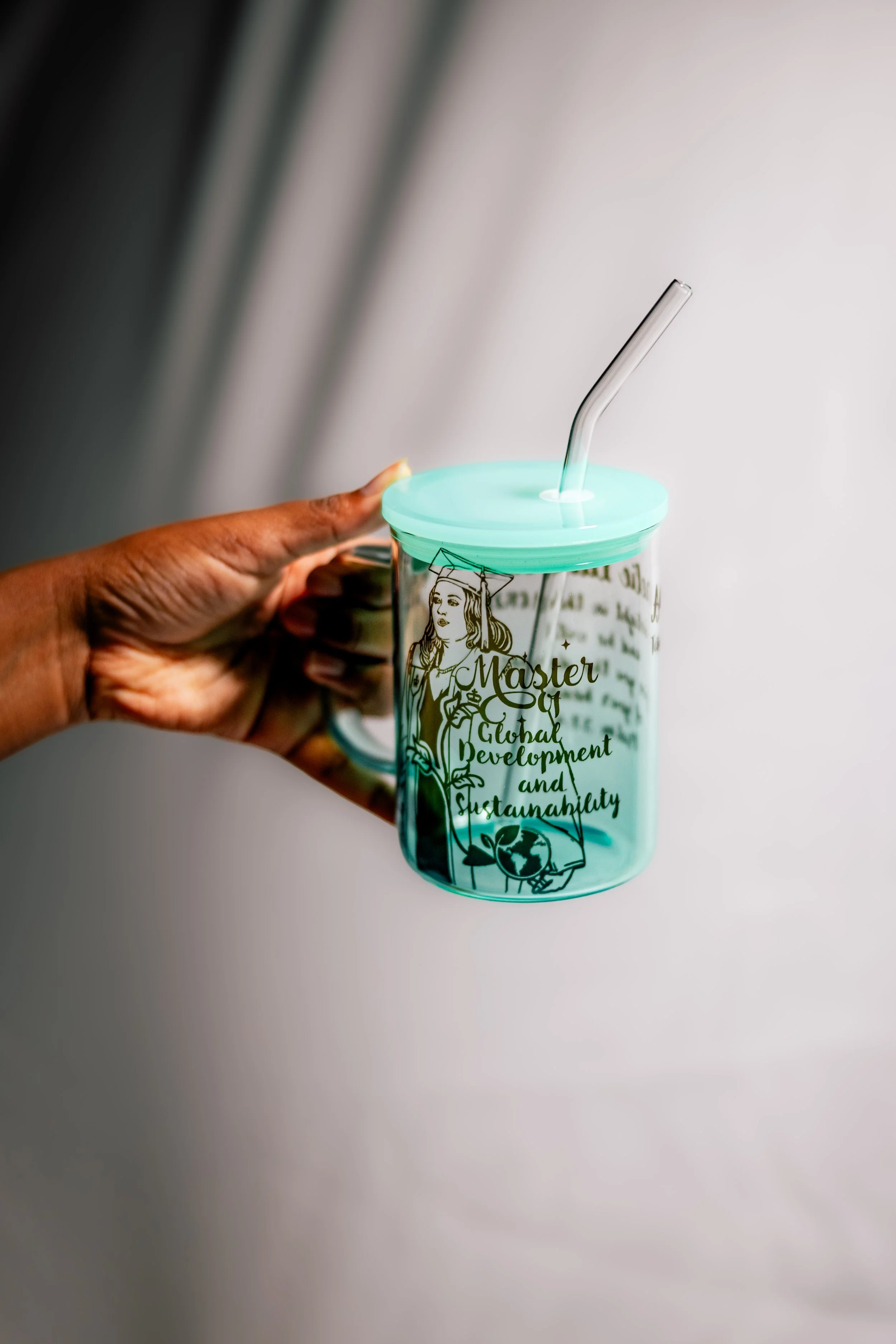 A hand holding a transparent tumbler with a mint green lid and a clear straw. The tumbler has a graphic of a woman with a crown and the words 'Master of Global Development and Sustainability.'