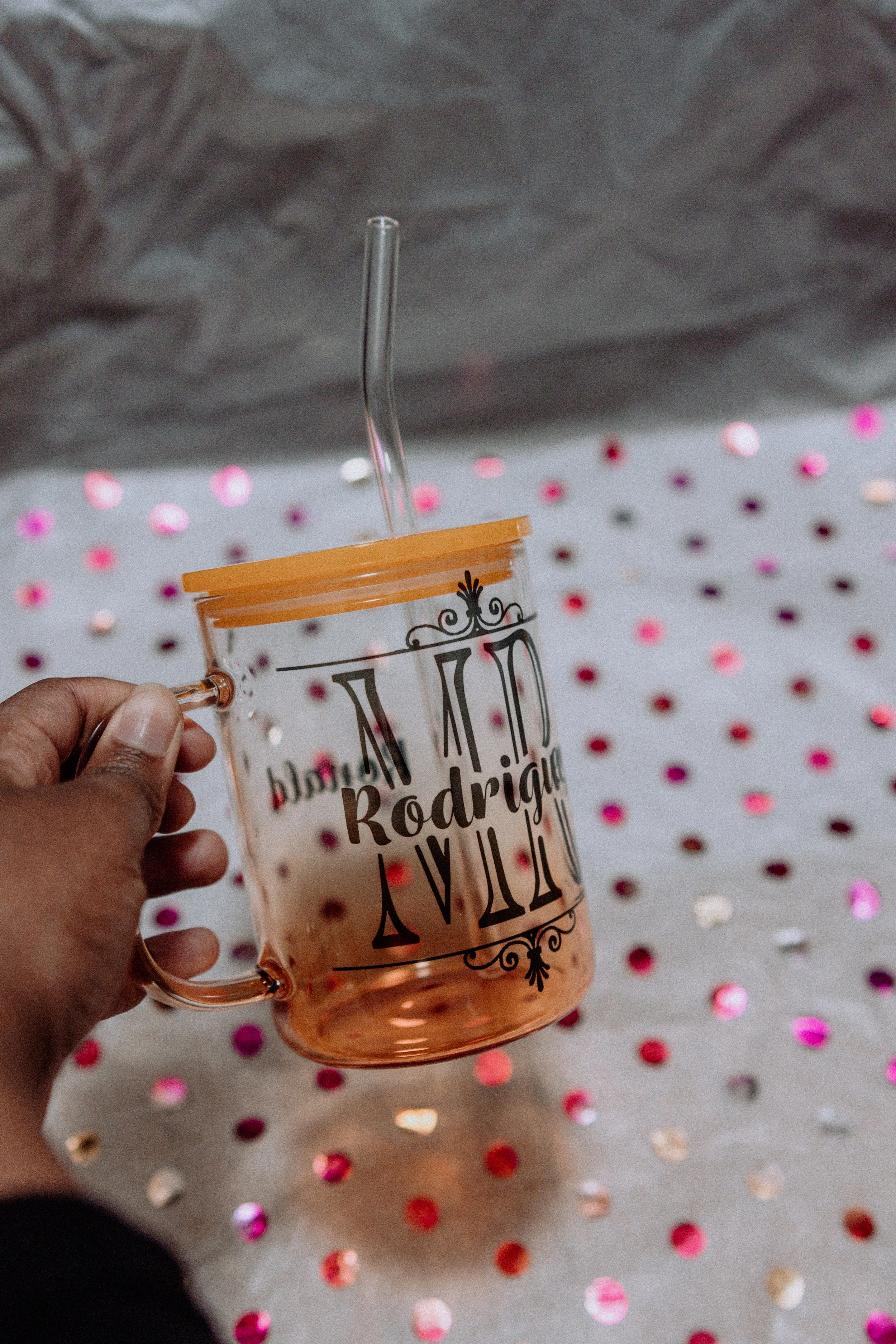 A person holding a transparent glass mug with black text with gold accents. The mug contains a pink liquid and a clear curved straw. The background has a white surface with multicolored shiny confetti dots.