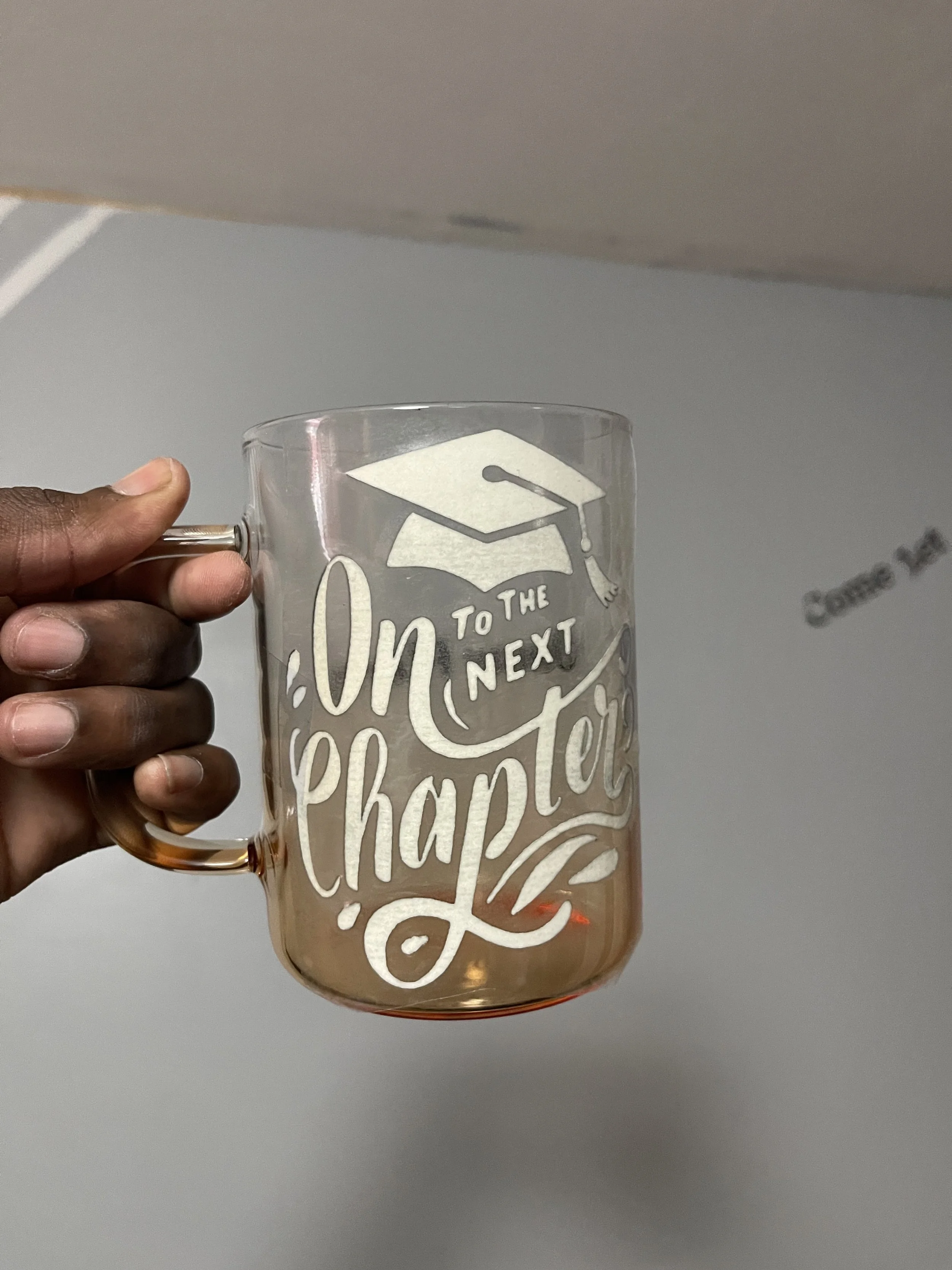 A hand holding a glass mug with white text and graphics about graduation and future plans, against a plain gray wall background.