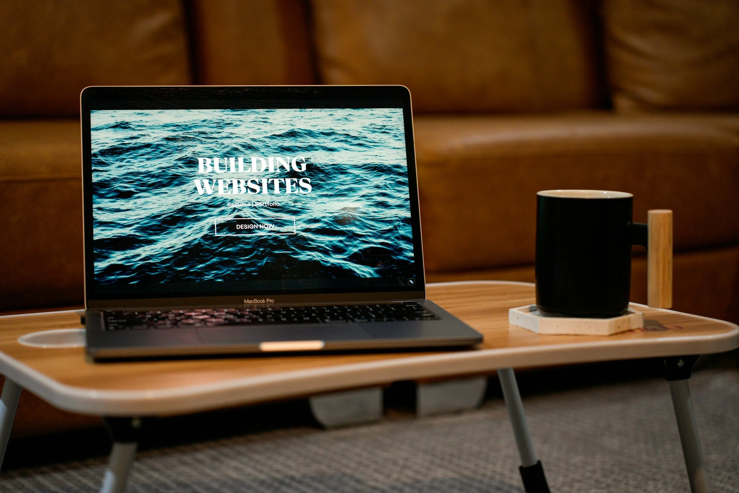 Laptop on a wooden table displaying a website with the text 'Building Websites' over a body of water background, next to a black mug on a coaster.