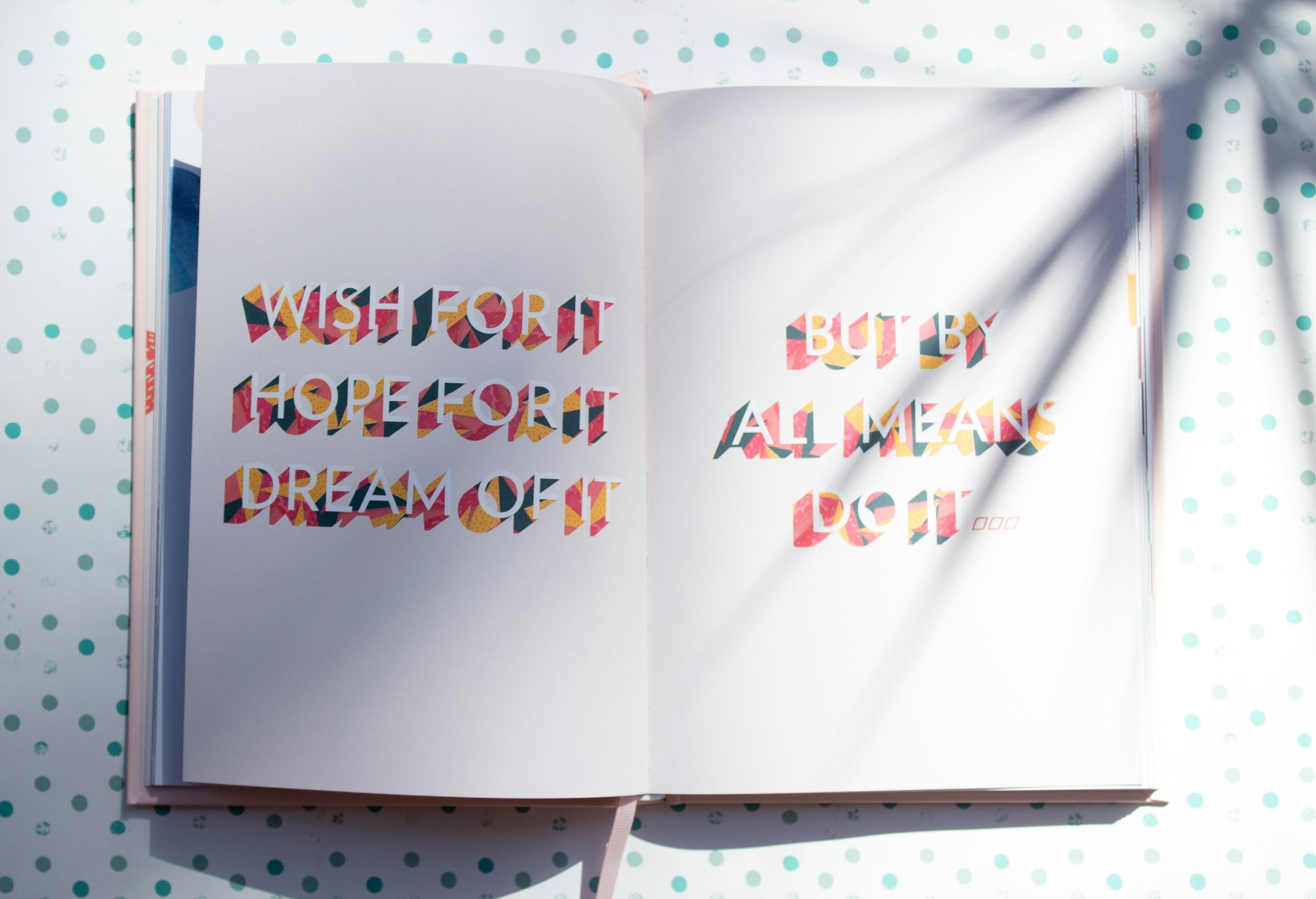 Open notebook on a polka dot surface with colorful, patterned text that reads: "Wish for it hope for it dream of it" on the left page and "but by all means do it" on the right page. Sunlight casting shadows across the pages.