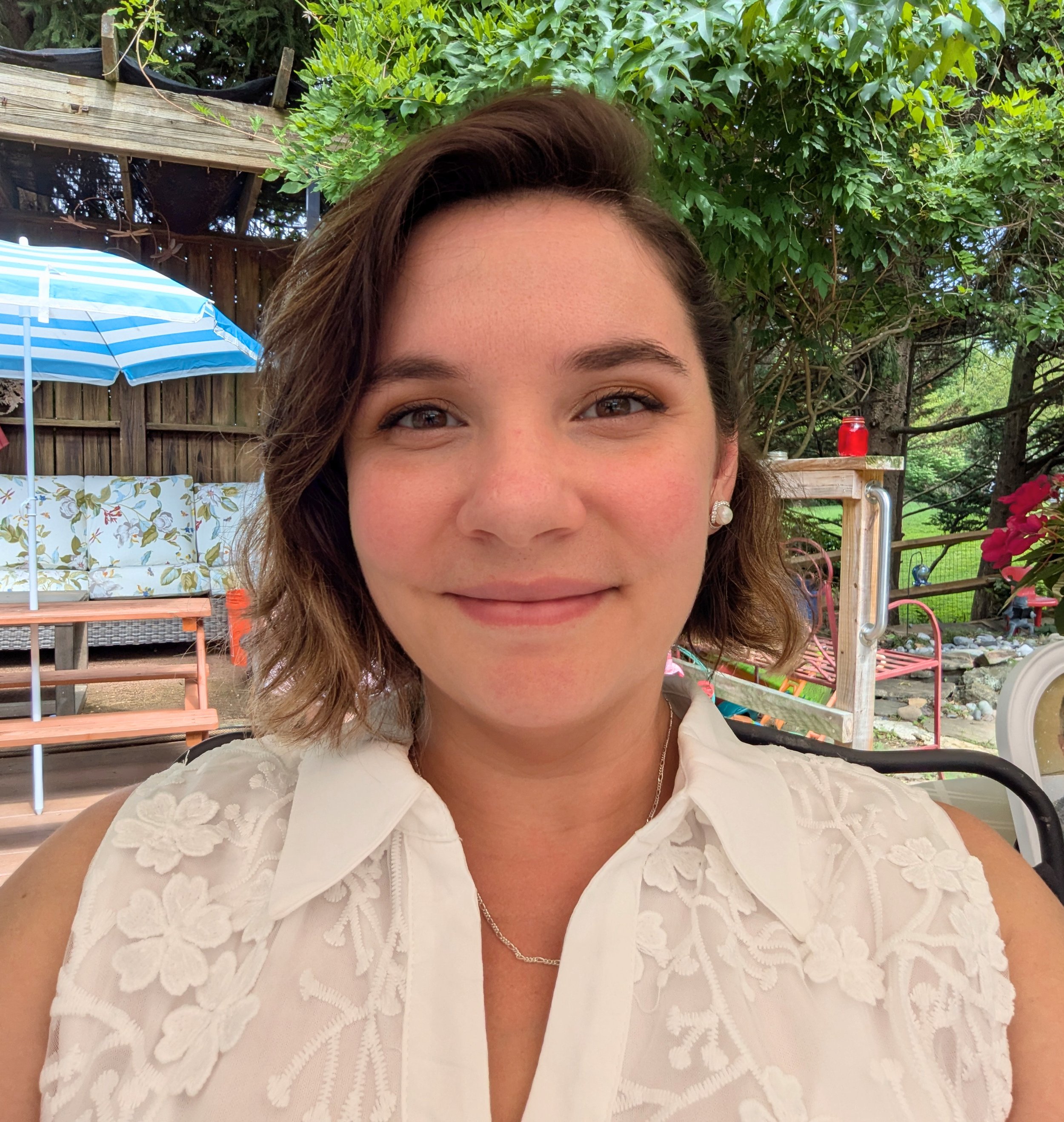 Close-up of Lyne Kolb, a woman with brown hair wearing a white embroidered blouse, smiling outdoors with green trees and patio furniture in the background.