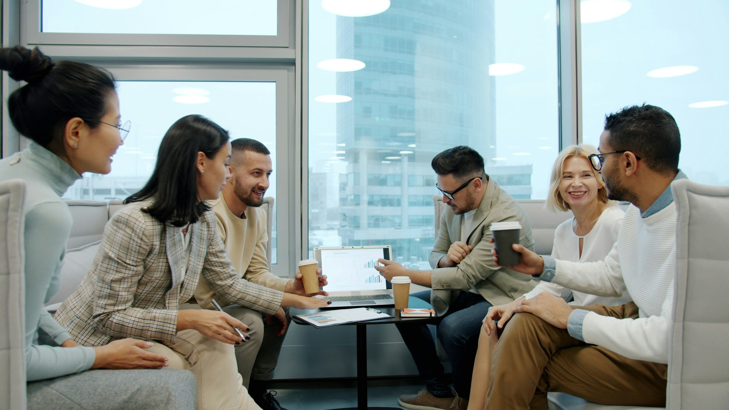 A diverse group of seven people gathered in a modern office lounge, engaged in conversation, with drinks in hand, next to a table with a laptop and documents, large windows with city skyscrapers in the background.