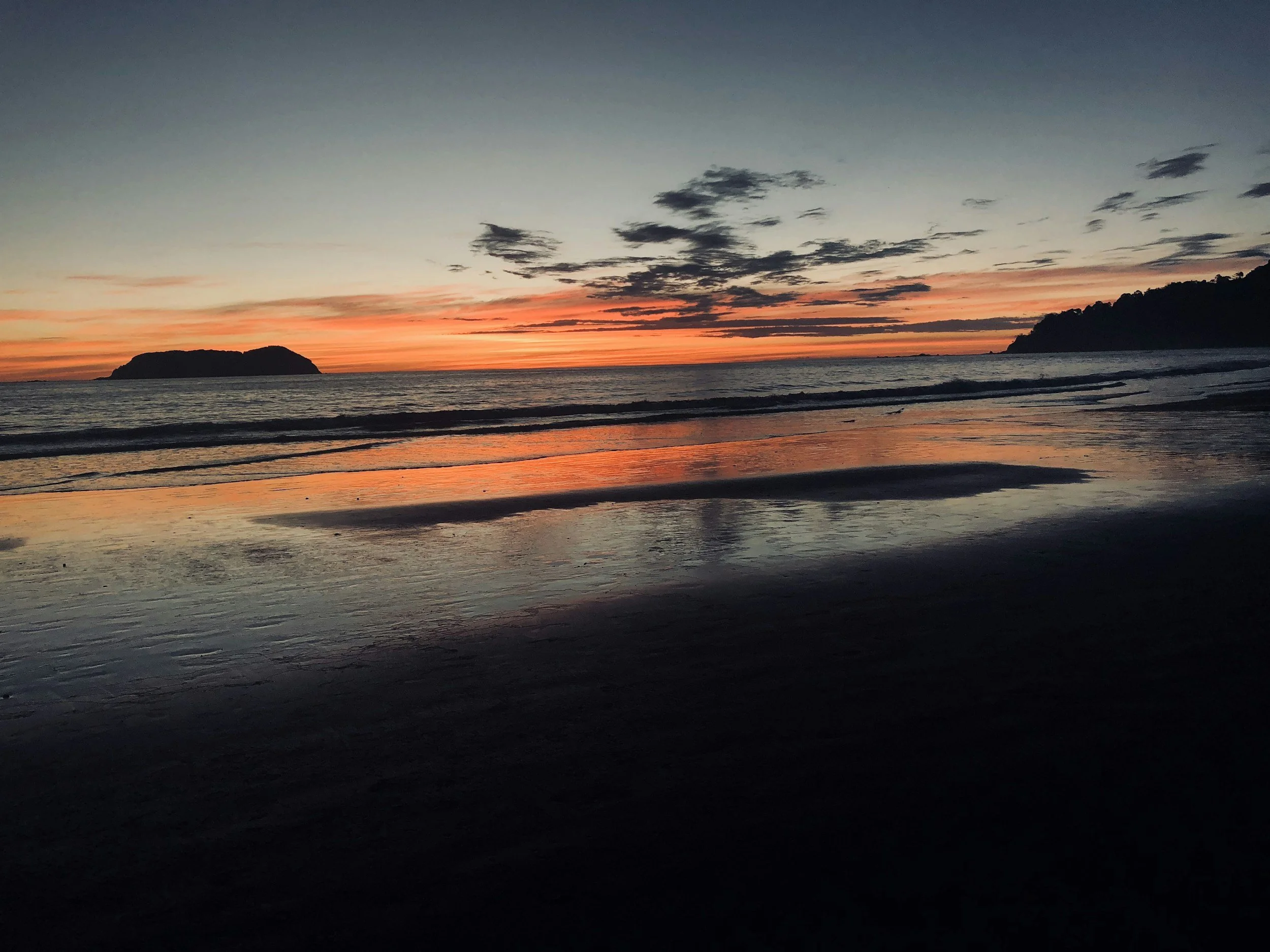 A sunset at the beach with an orange and pink sky, calm water reflecting the colors, and a silhouetted island on the horizon.