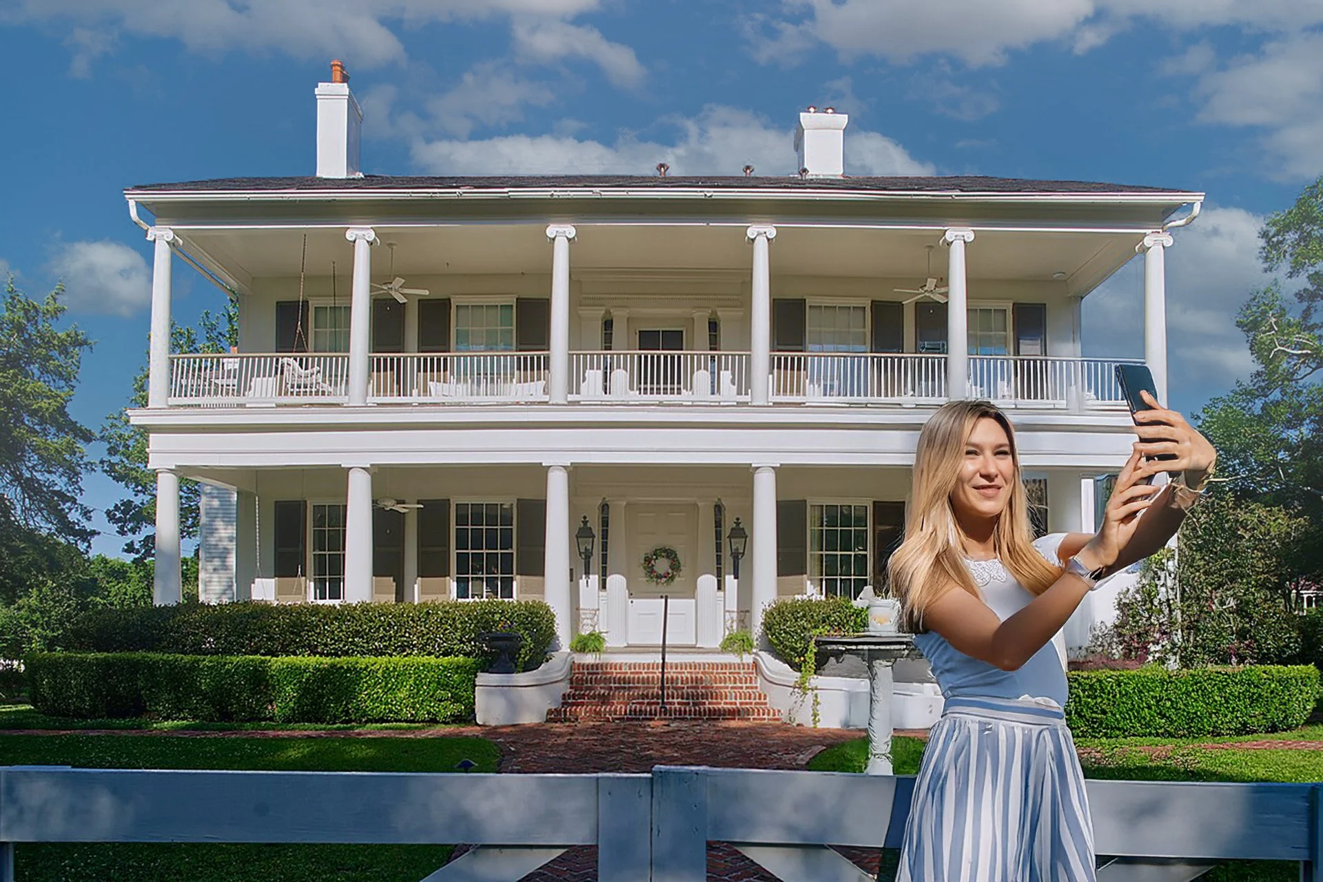 A woman with blonde hair, wearing a white shirt and striped pants, takes a selfie in front of a large, white, two-story house with columns and a wraparound porch on a sunny day.