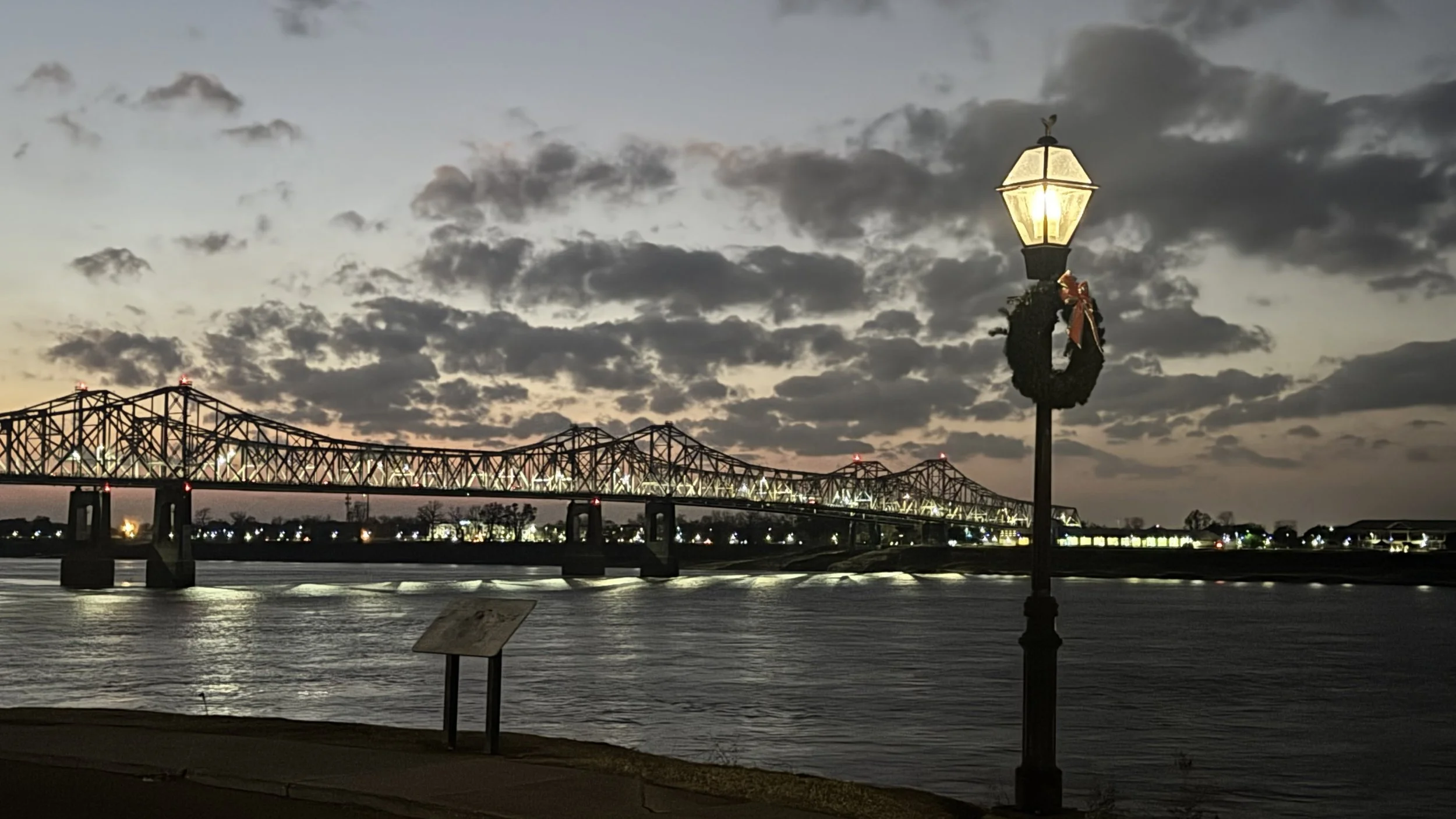 A scenic view of a bridge over a river during twilight with a decorated lamppost in the foreground and a cloudy sky.