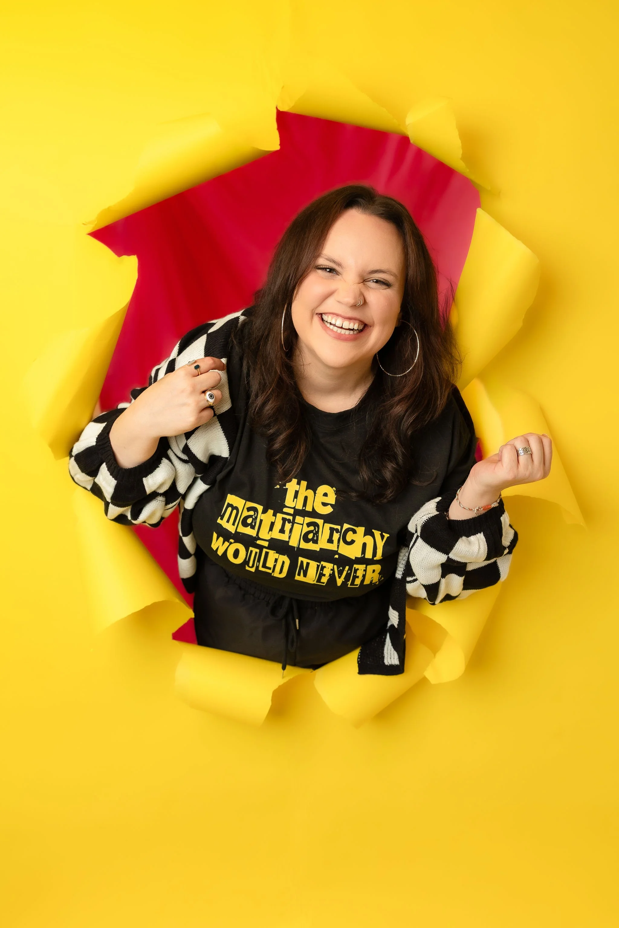 A woman smiling, with a yellow background. She is wearing glasses, a plaid shirt, and a graphic t-shirt.