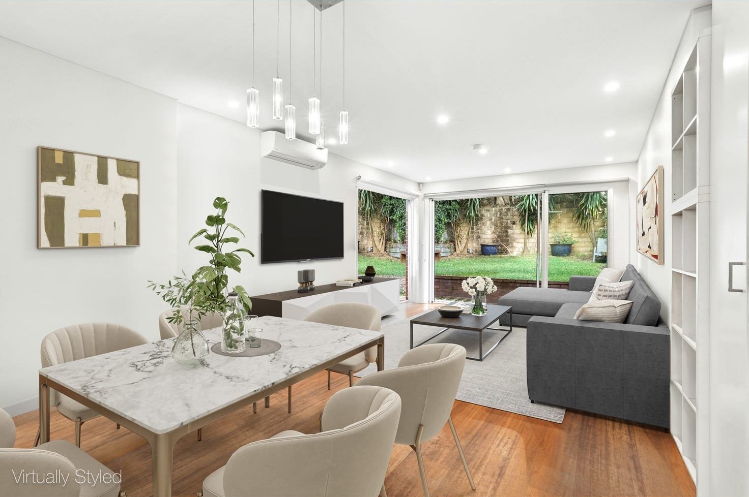 Modern living room with a marble dining table, beige chairs, gray sectional sofa, wall-mounted TV, sliding glass doors leading to a backyard with grass and trees, indoor plants, abstract art, and wooden flooring.