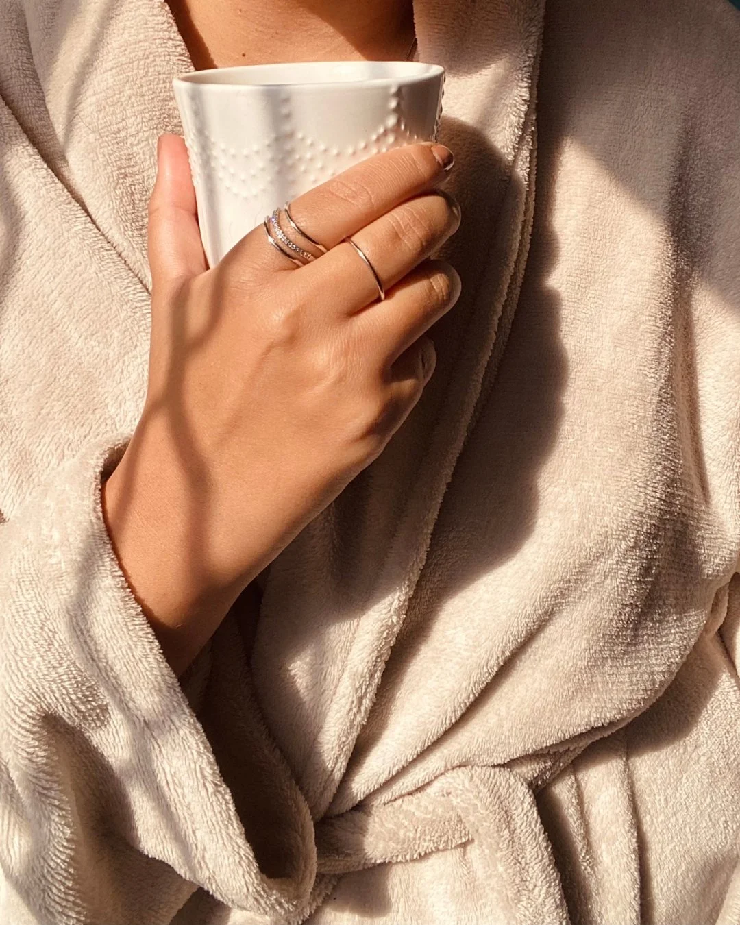 Person holding white ceramic mug with textured pattern while wearing multiple rings on their left hand, dressed in a beige plush robe.
