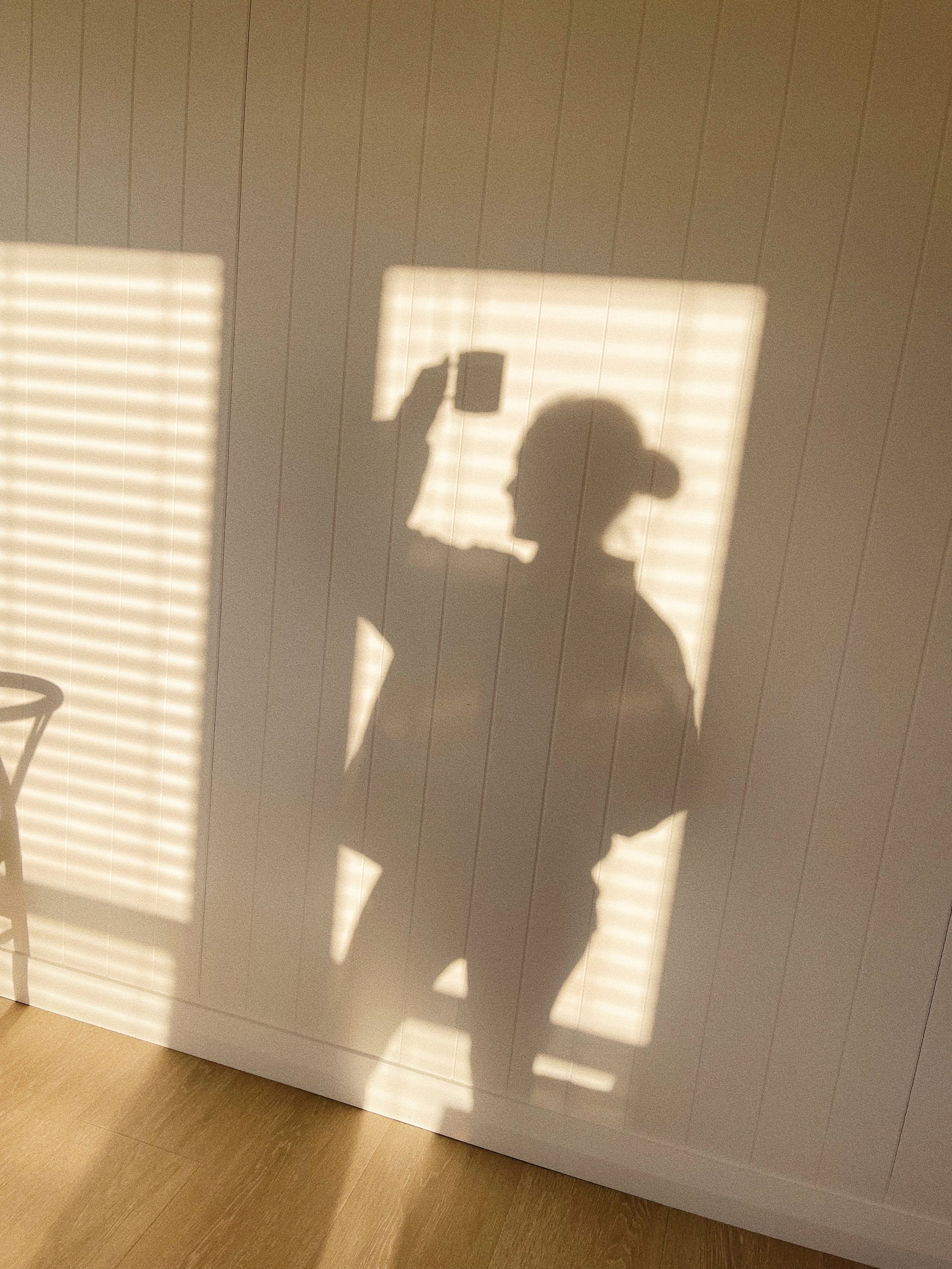 The shadow of a woman with glasses and a bun, holding a phone, cast on a wall with window blinds letting in sunlight.