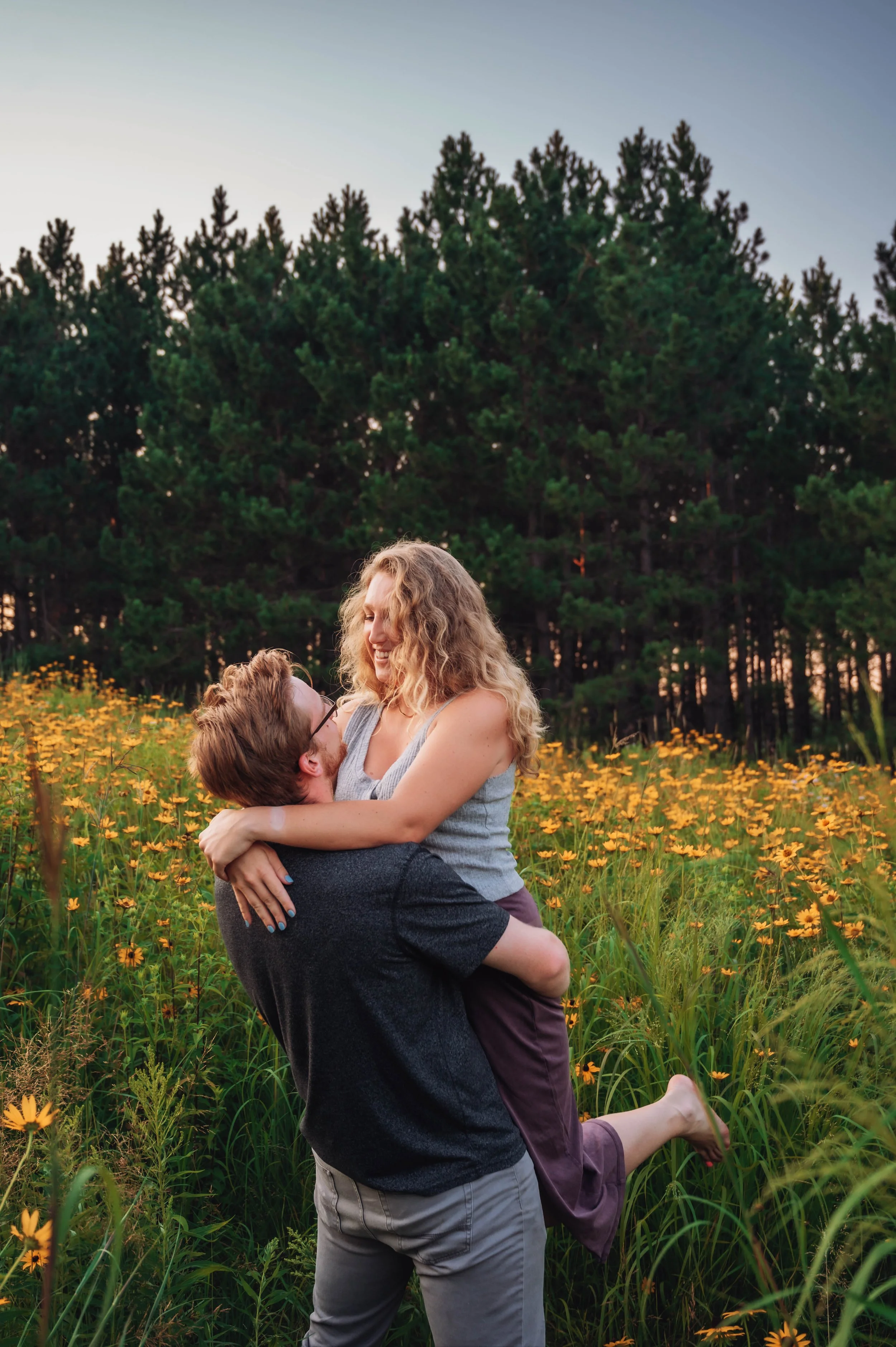 A man lifting a woman in a field of yellow flowers with a forest in the background during sunset, both smiling and looking at each other.
