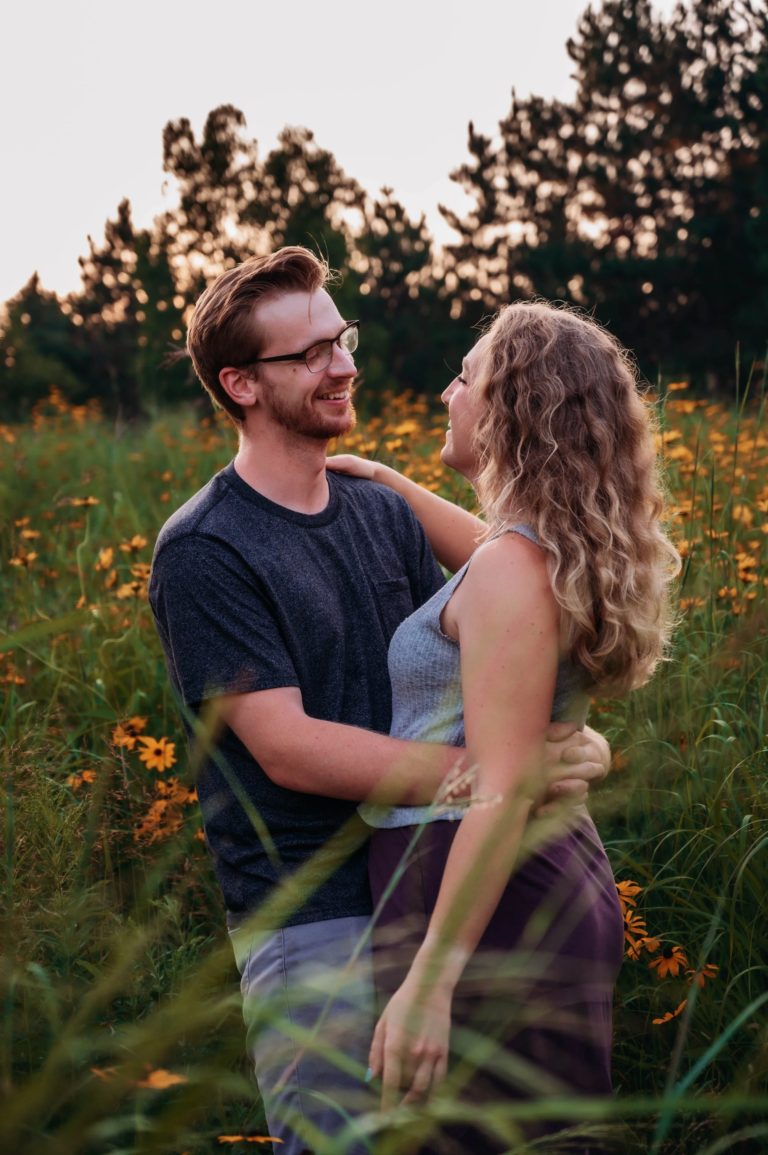 A young couple smiling and embracing in a field of yellow flowers during sunset, with trees in the background.
