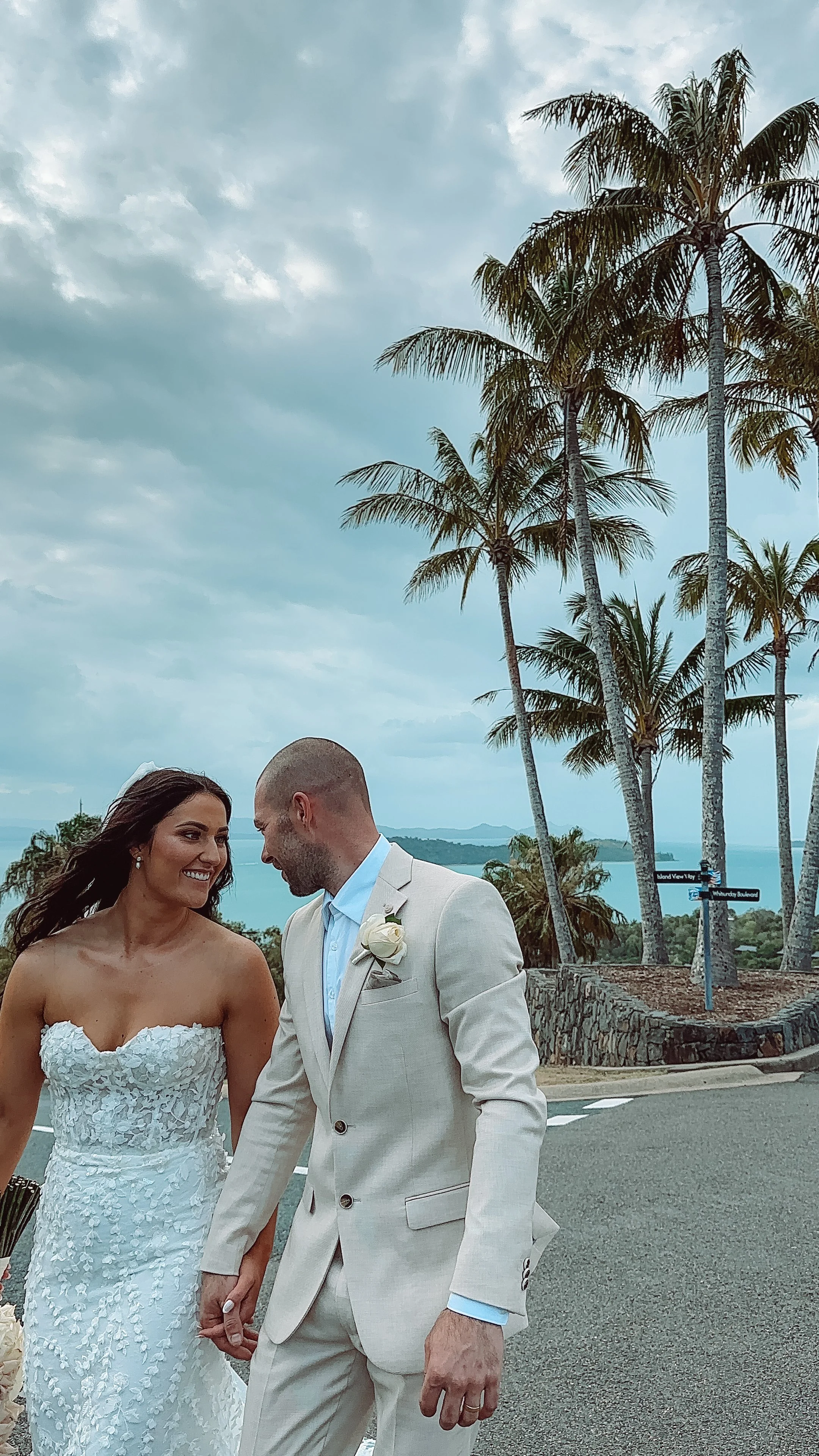 A bride and groom holding hands and smiling at each other outdoors with palm trees and a body of water in the background.