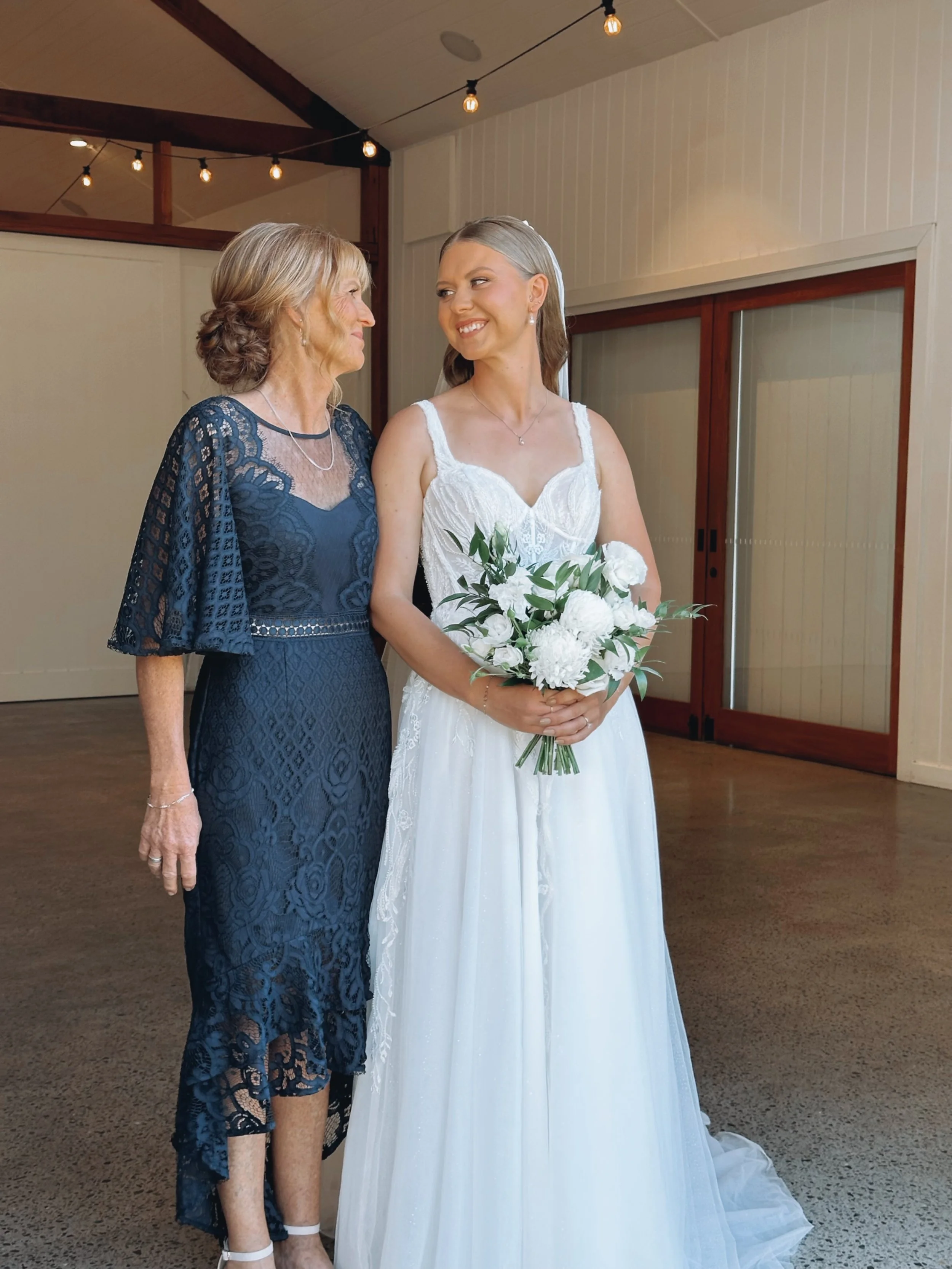 A young woman in a white wedding dress holding a bouquet of white flowers standing next to an older woman in a blue lace dress.