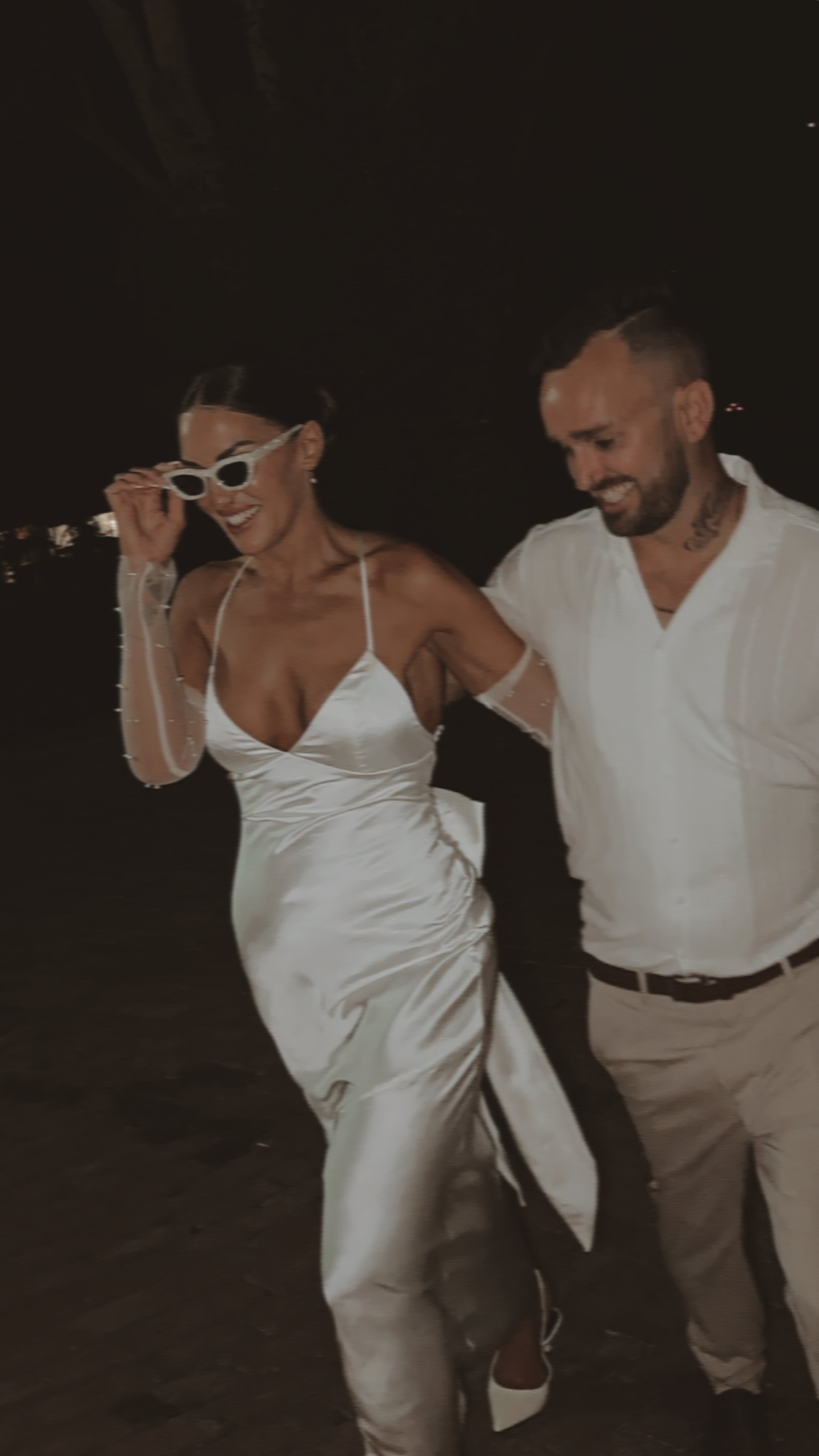 A woman in a white satin dress with sunglasses on her head is holding hands with a man in a white shirt and beige pants. They are smiling and walking together at night.