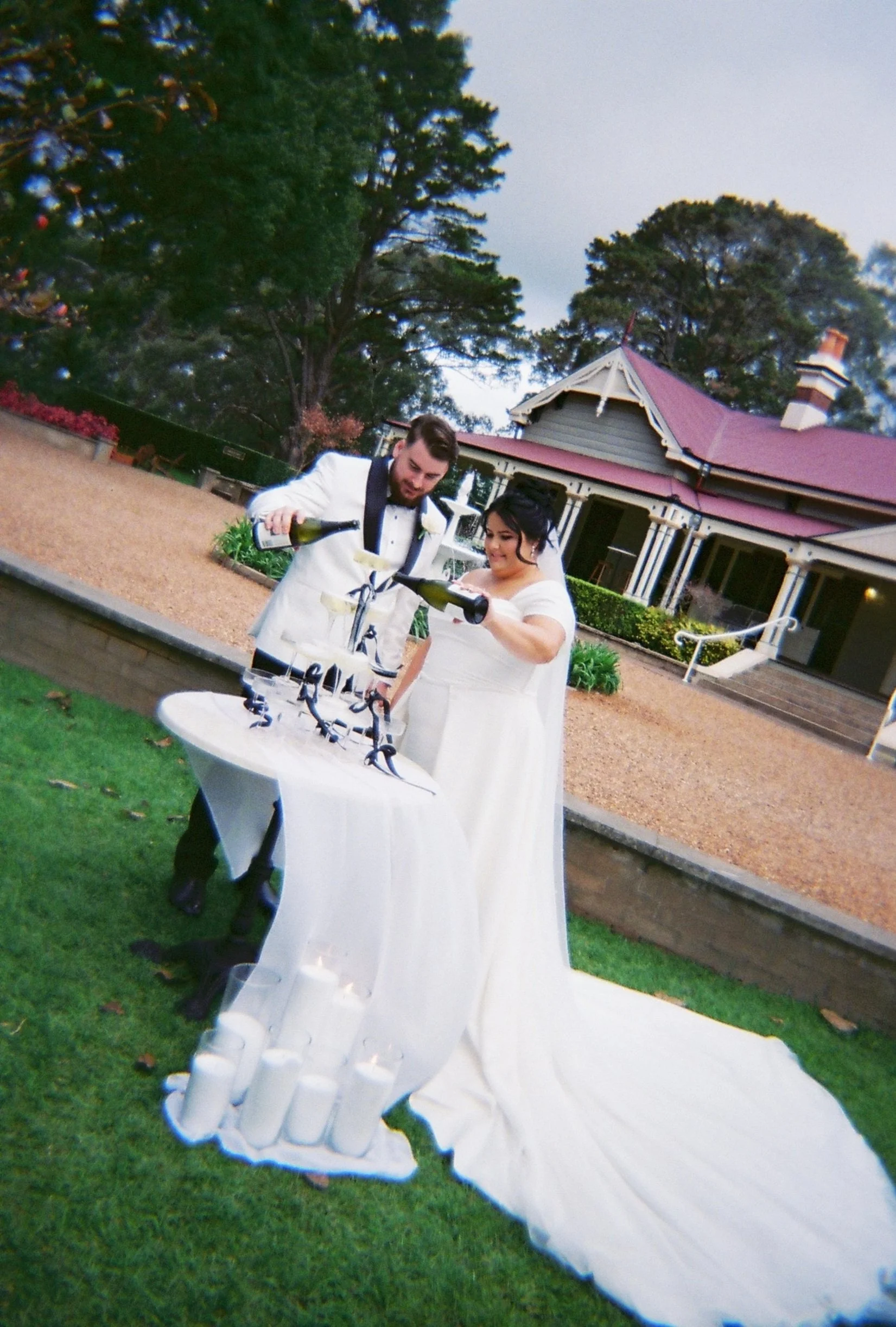 A newlywed couple in wedding attire pouring champagne into glasses at an outdoor reception, with a white table, candles, and a vintage house in the background.