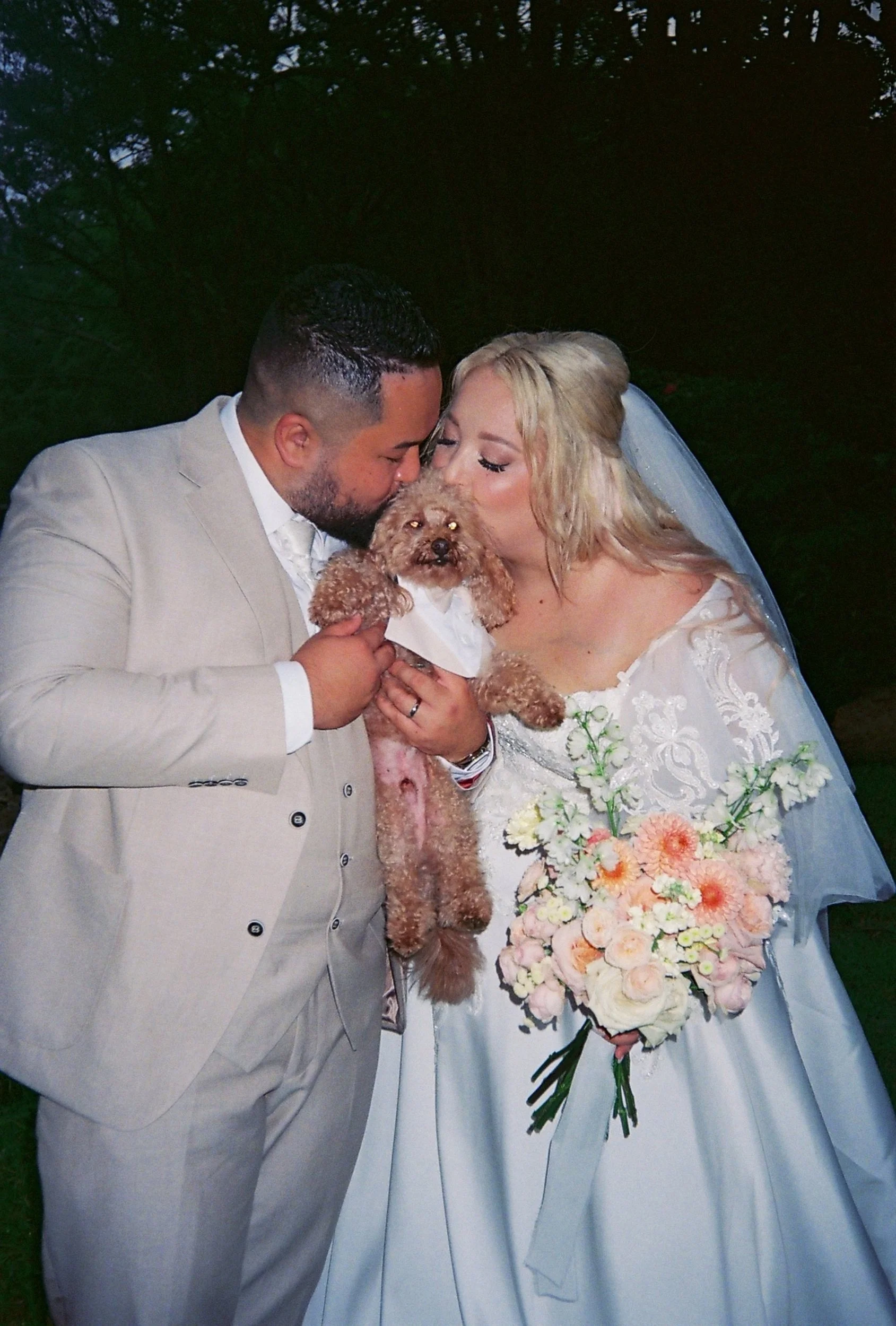 A bride and groom kiss a small dog at their wedding, holding a bouquet of flowers. The groom is wearing a light-colored suit, and the bride is in a white wedding dress with lace details and a veil. The scene is outdoors with trees in the background.