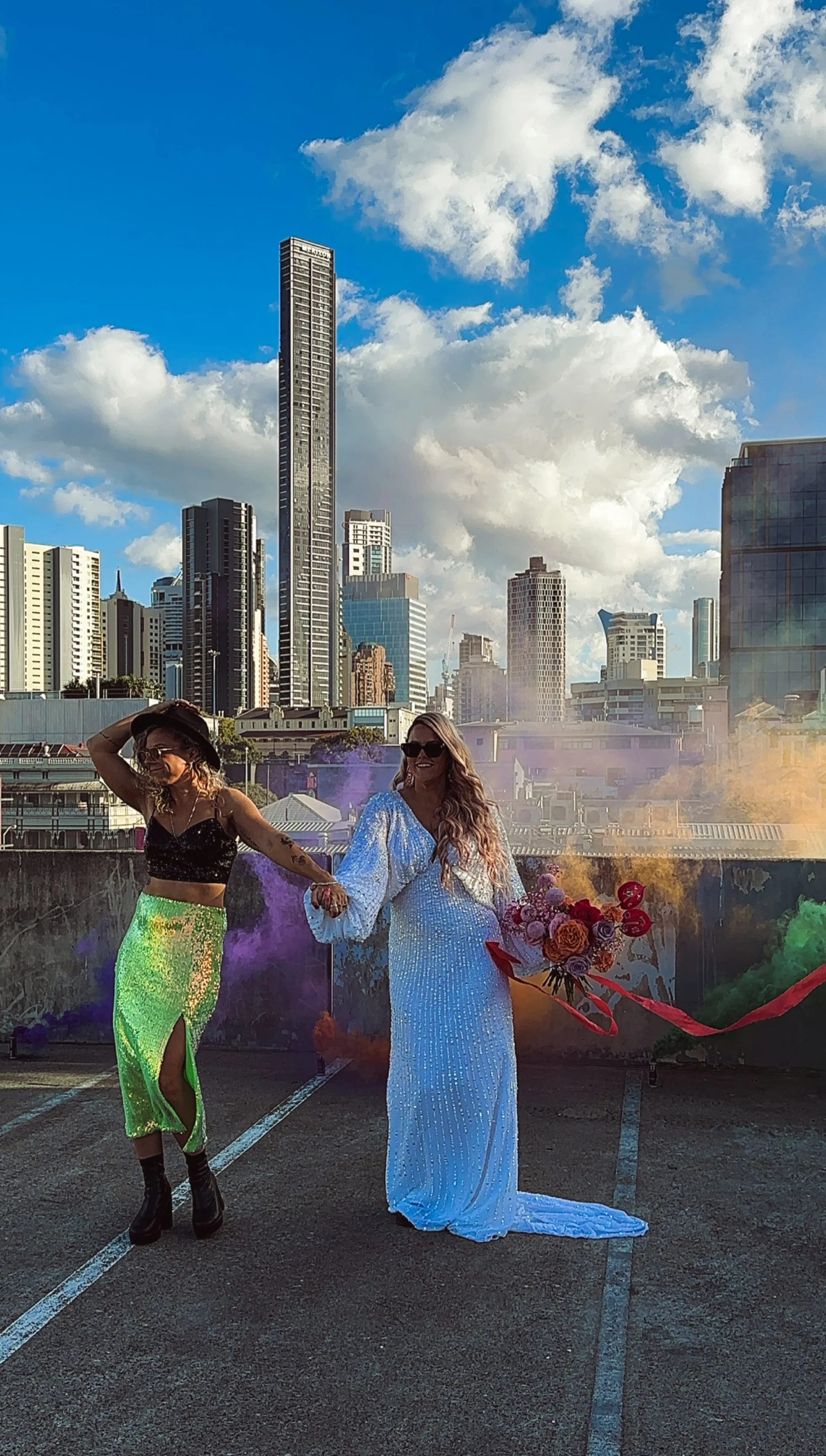Two women holding hands in a parking lot with a city skyline in the background. One is wearing a black top and a green sequin skirt, and the other is wearing a white dress and holding a bouquet of flowers. Colorful smoke is in the air.