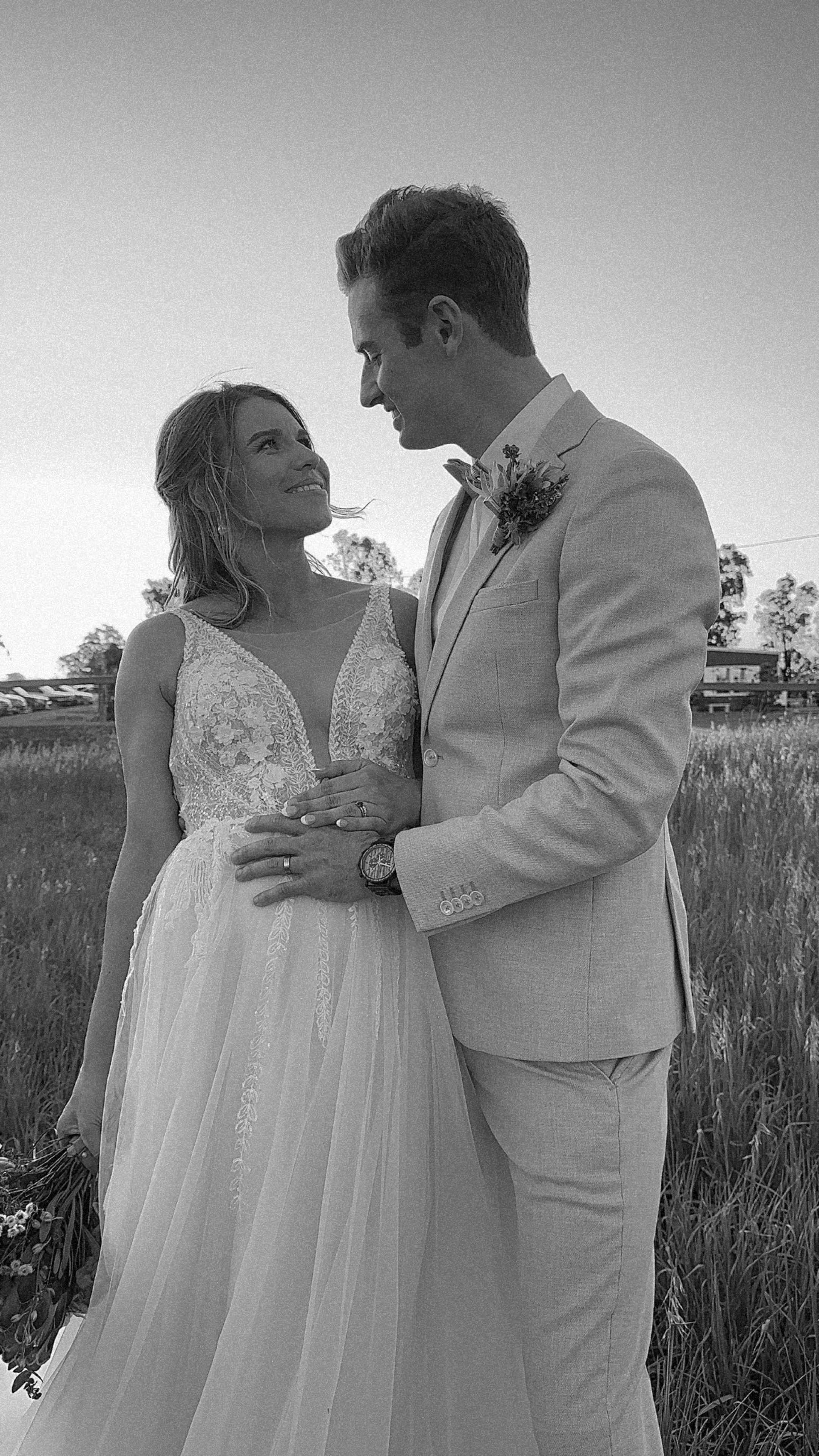 A black and white photo of a groom and bride on their wedding day, standing outdoors in a grassy field, smiling at each other.