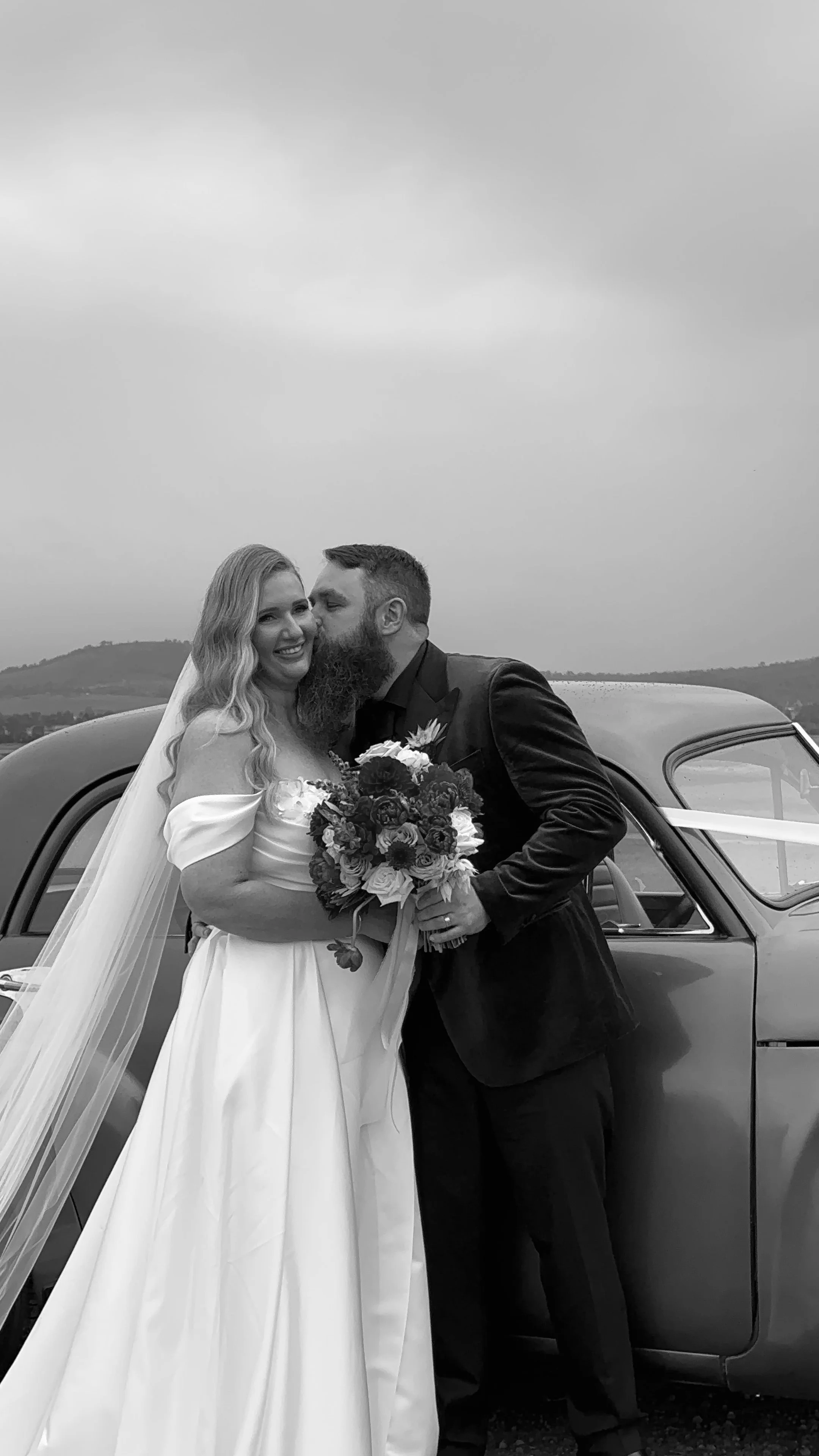 A newlywed couple in wedding attire standing by a vintage car outdoors, with the groom kissing the bride on her cheek, holding a bouquet of flowers, on a cloudy day.