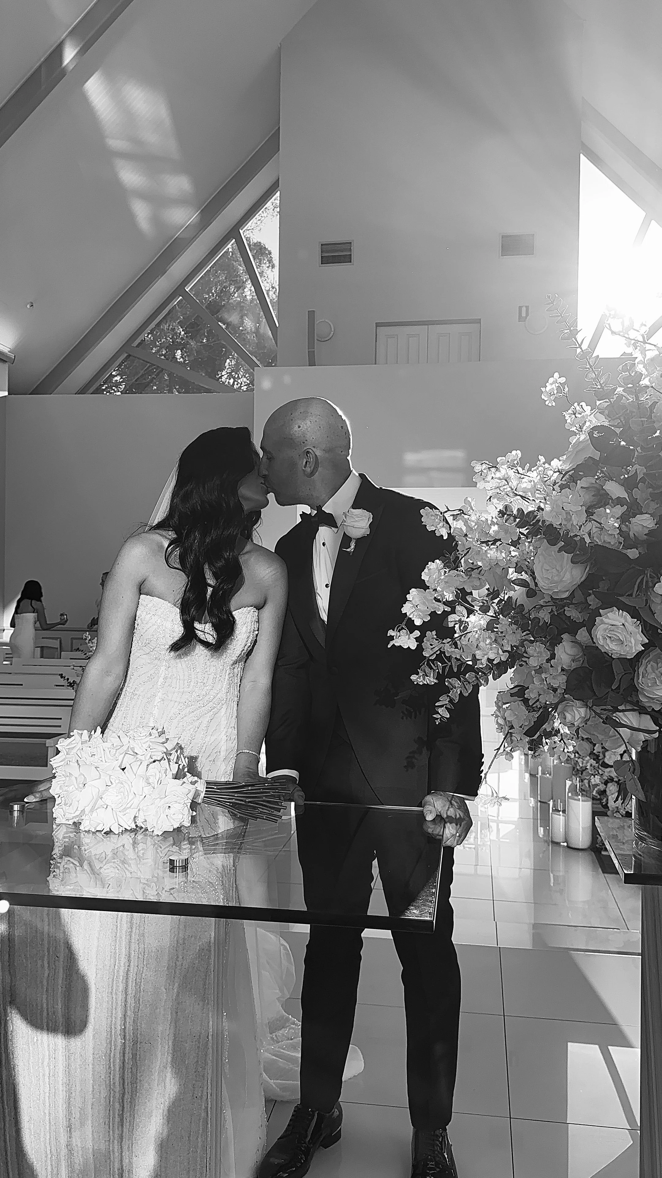 Black-and-white photo of a bride and groom sharing a kiss in a church, with the bride in a strapless wedding dress holding a bouquet of white roses, and the groom in a black tuxedo with a boutonnière, standing next to a large floral arrangement.