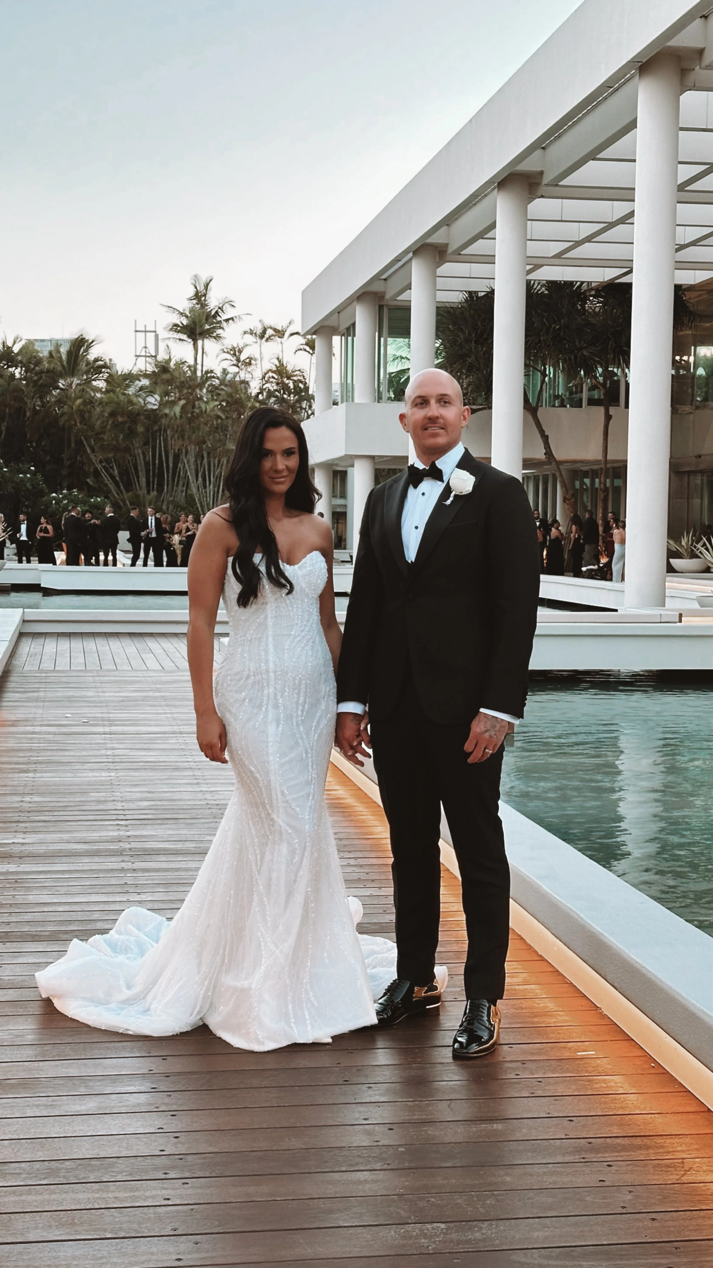 A bride and groom standing outdoors on a wooden deck near a pool, holding hands and posing for a photo during their wedding celebration at a modern venue with glass walls and palm trees in the background.