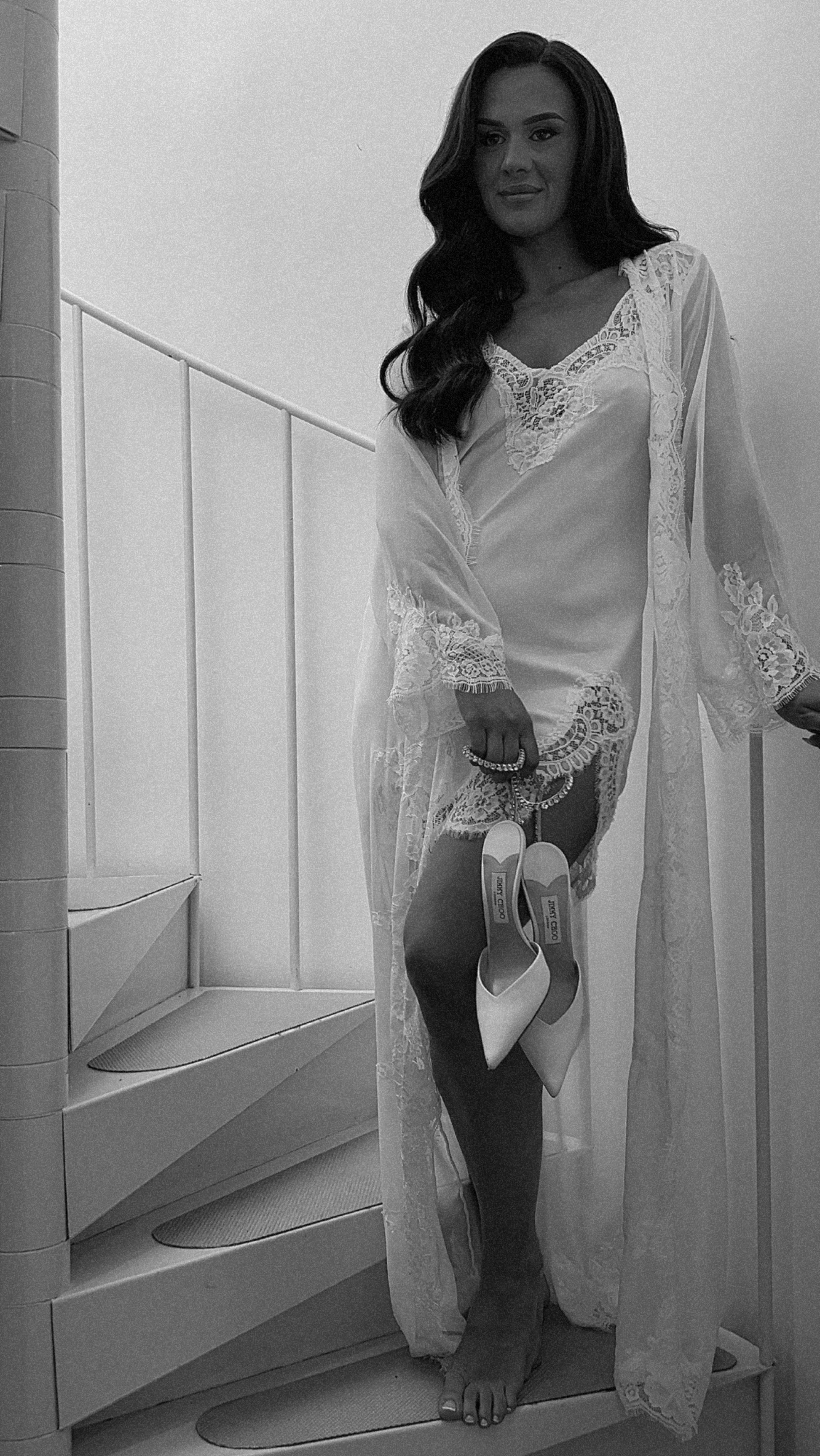 A woman in lingerie and a sheer robe standing on a staircase, holding a pair of high heel shoes and a beaded necklace.