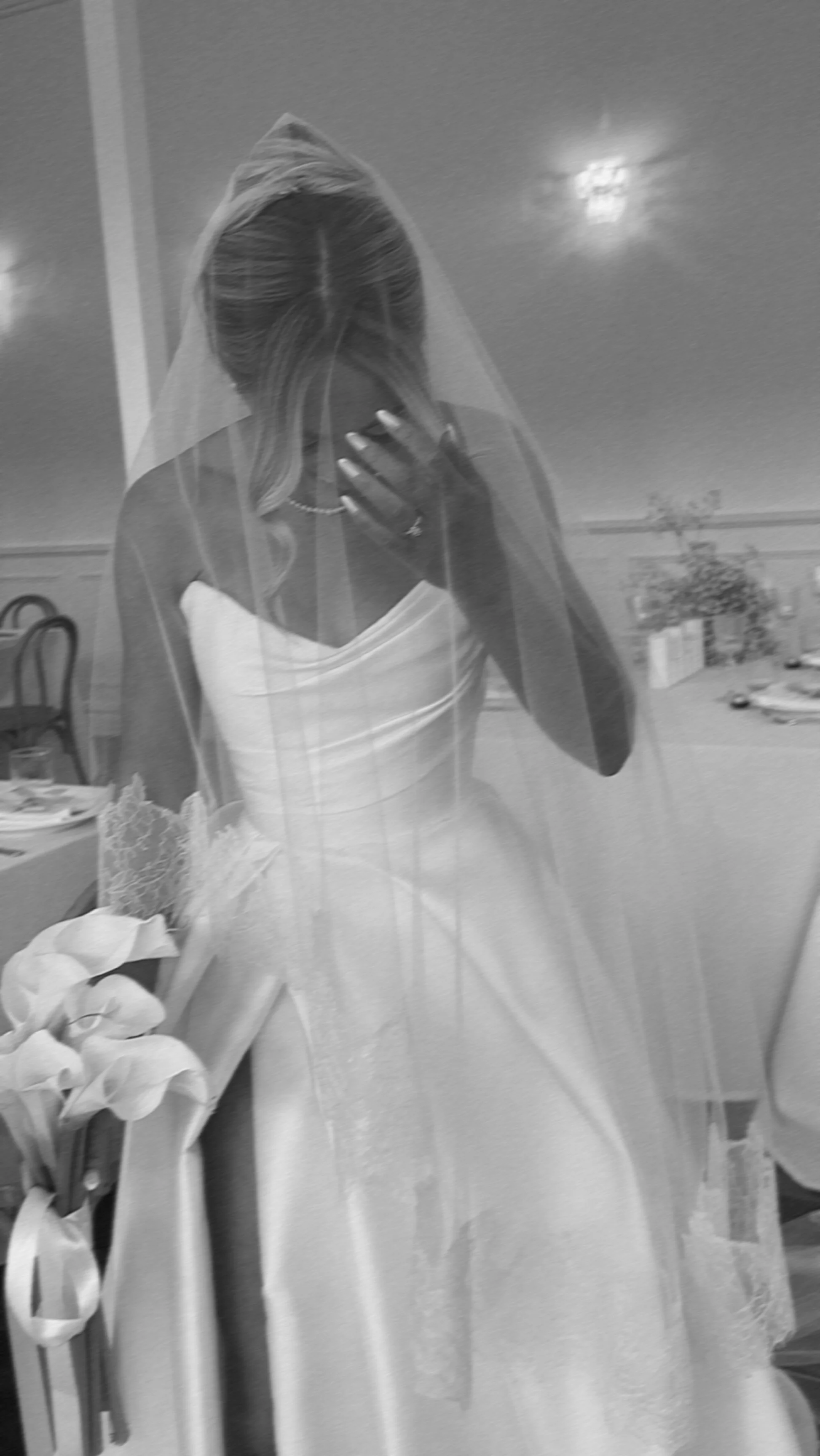 A woman in a wedding dress and veil taking a mirror selfie, with her face covered by her hand.