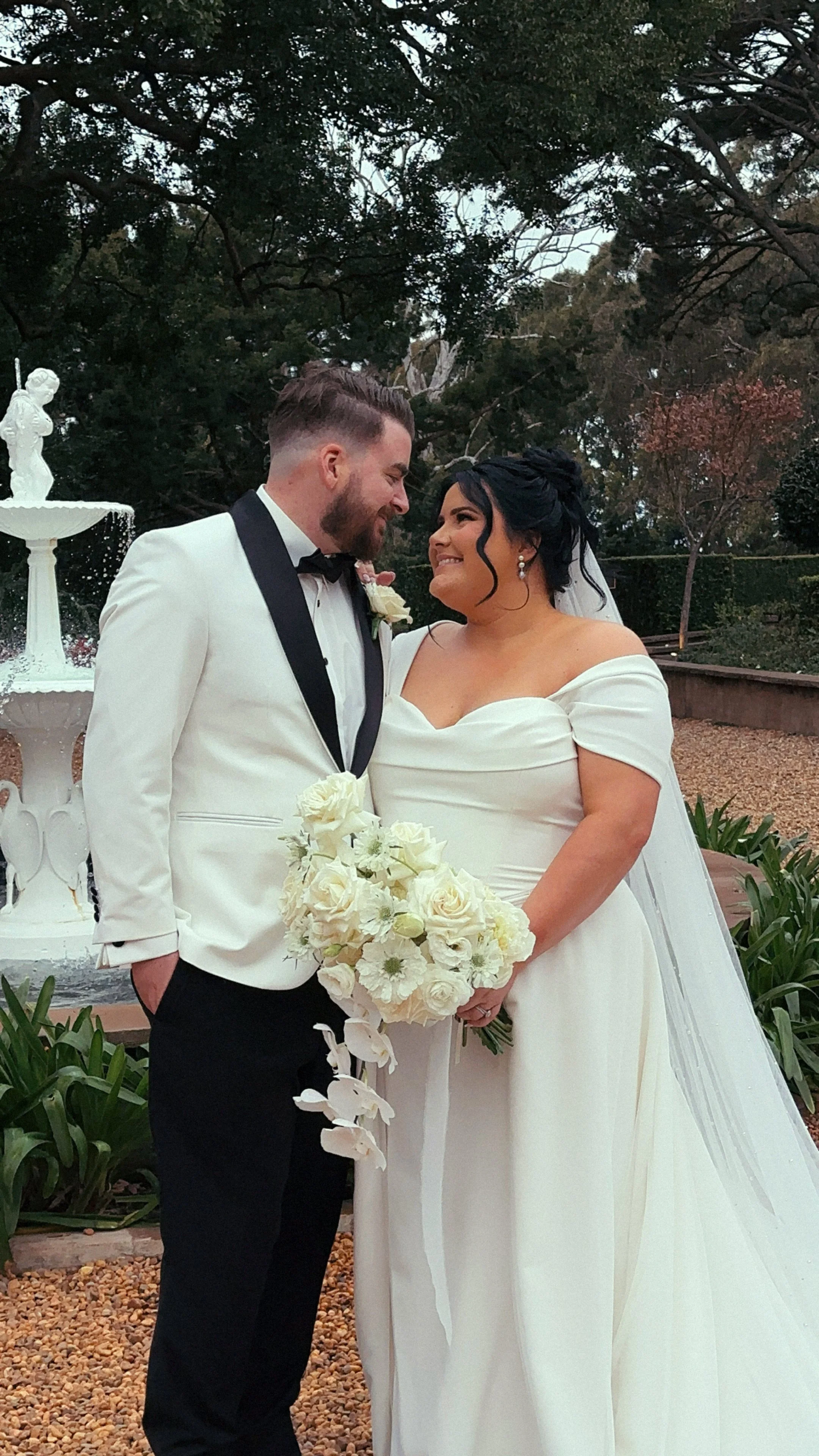 A newlywed couple dressed in wedding attire, standing outdoors near a fountain, smiling and gazing at each other. The bride is holding a bouquet of white flowers.