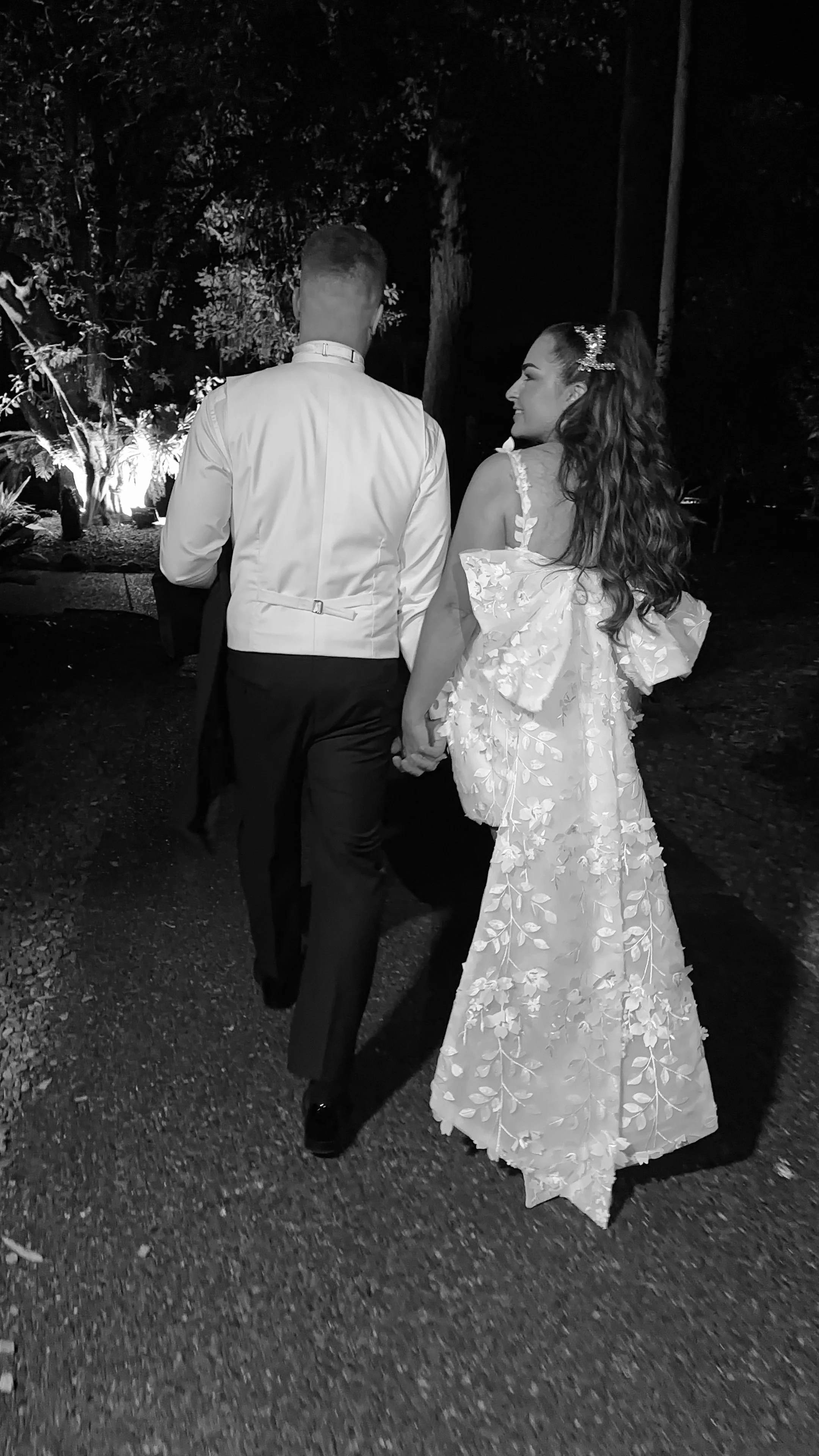 A black and white photo of a newlywed couple holding hands and walking together at night, with an outdoor setting and illuminated trees in the background.