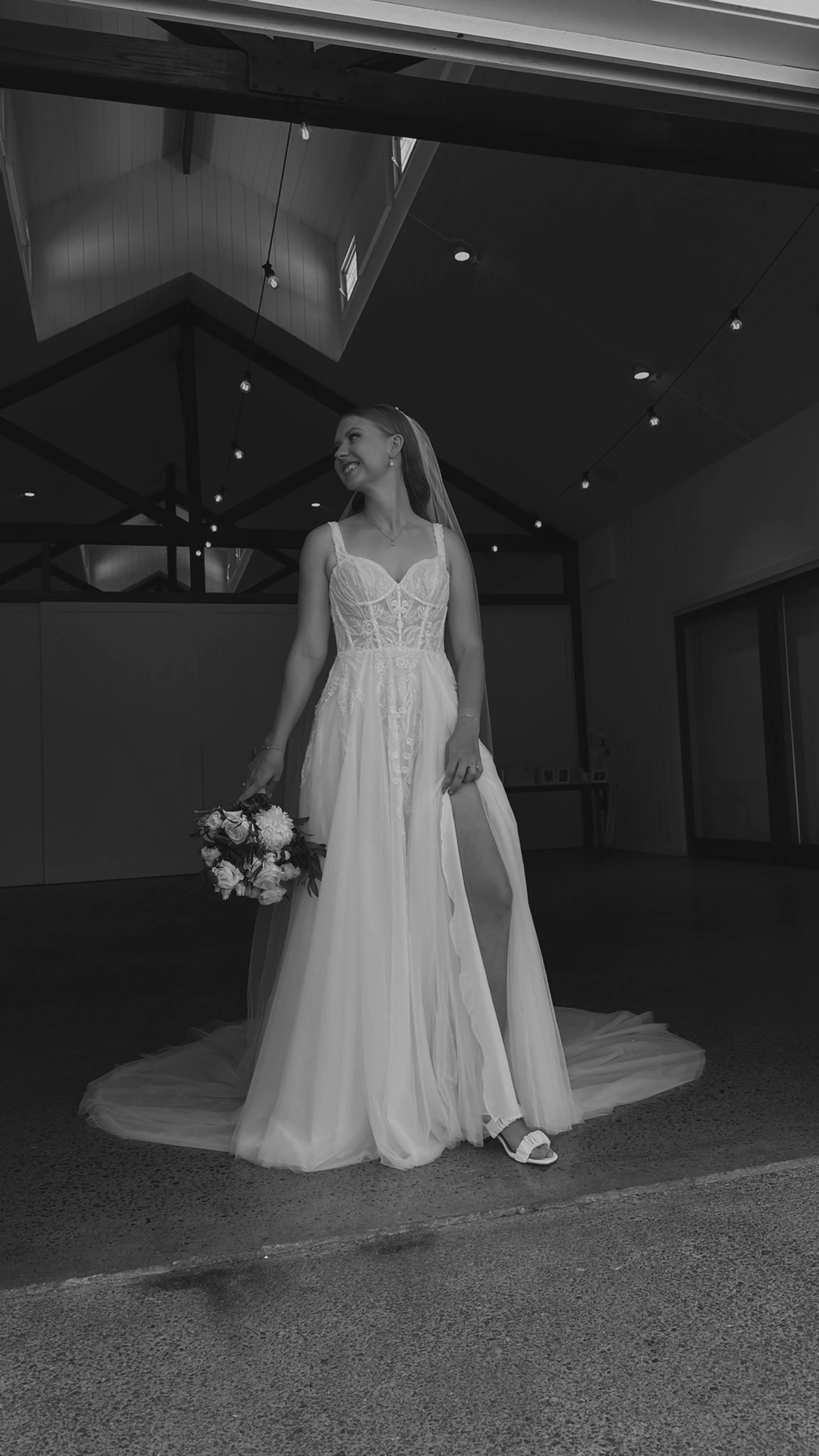 Black and white photo of a smiling bride holding a bouquet of flowers, wearing a wedding dress with a slit, inside a modern building with high ceilings and string lights.