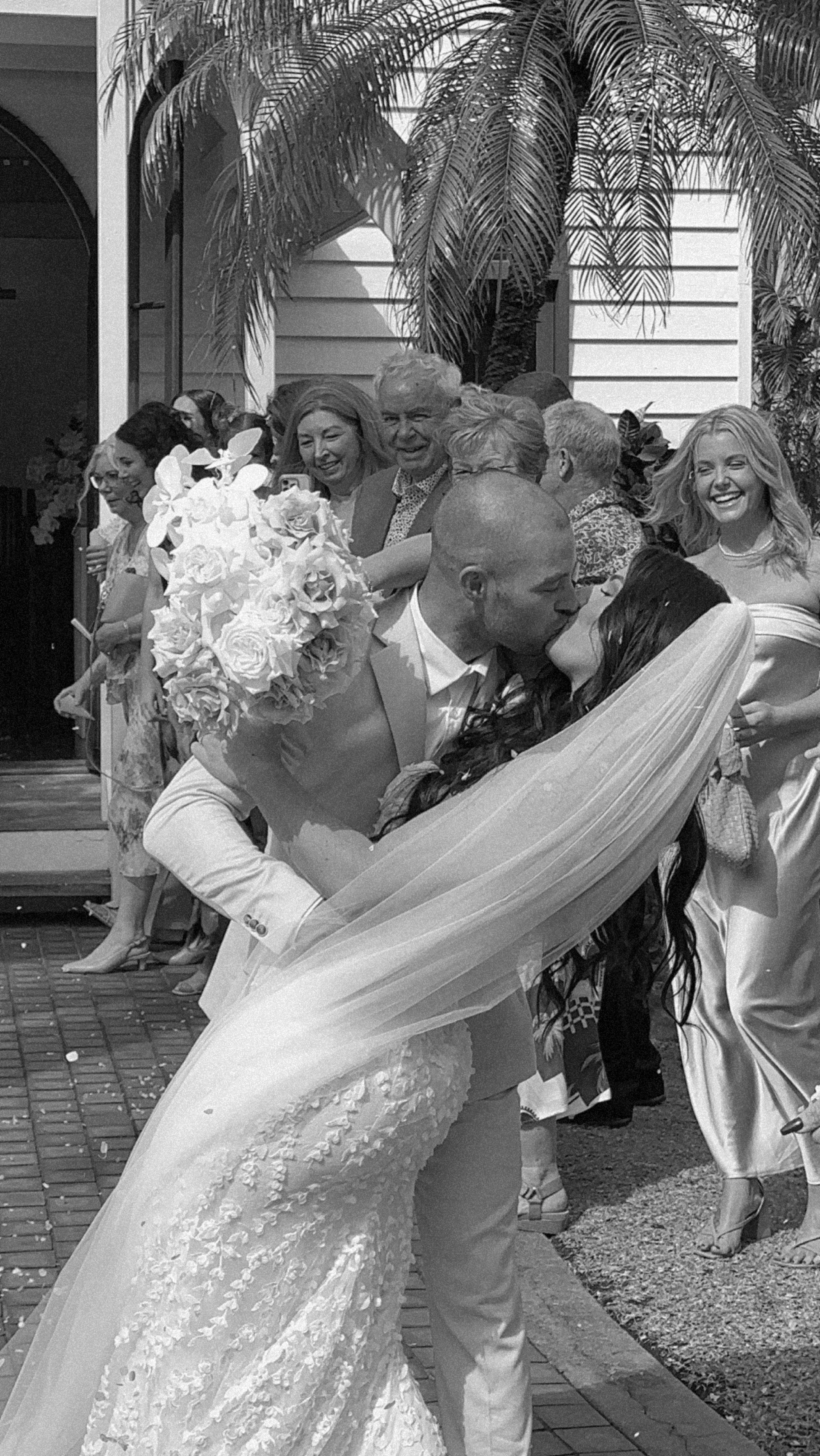 A black and white photo of a wedding scene with newlywed couple kissing, surrounded by friends and family. The bride wears a lace wedding gown with a veil, and the groom is in a light-colored suit holding a bouquet of flowers. People in the backgroun