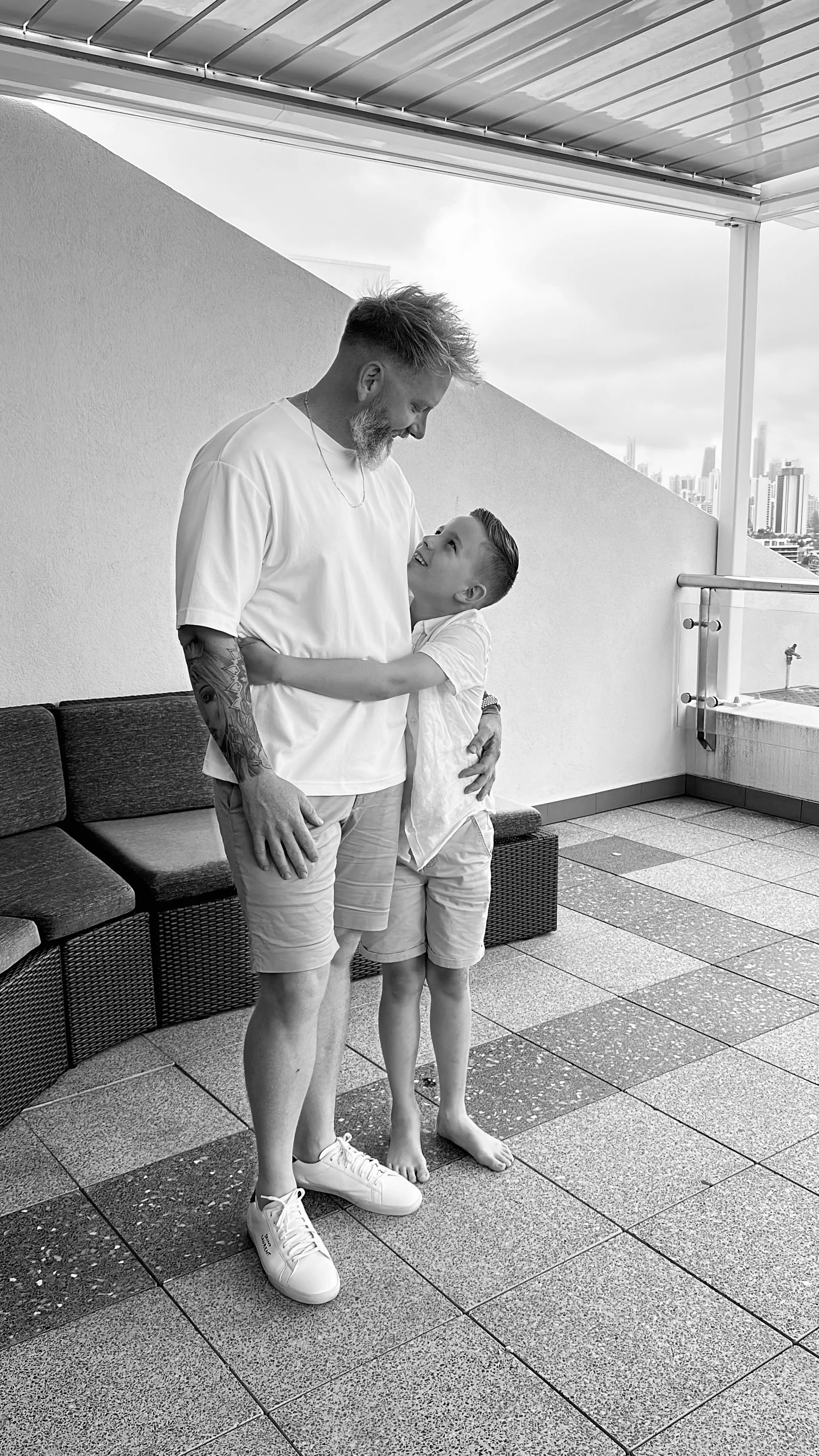 A man and a boy sharing a hug on an outdoor balcony with a city skyline in the background, black and white photo.