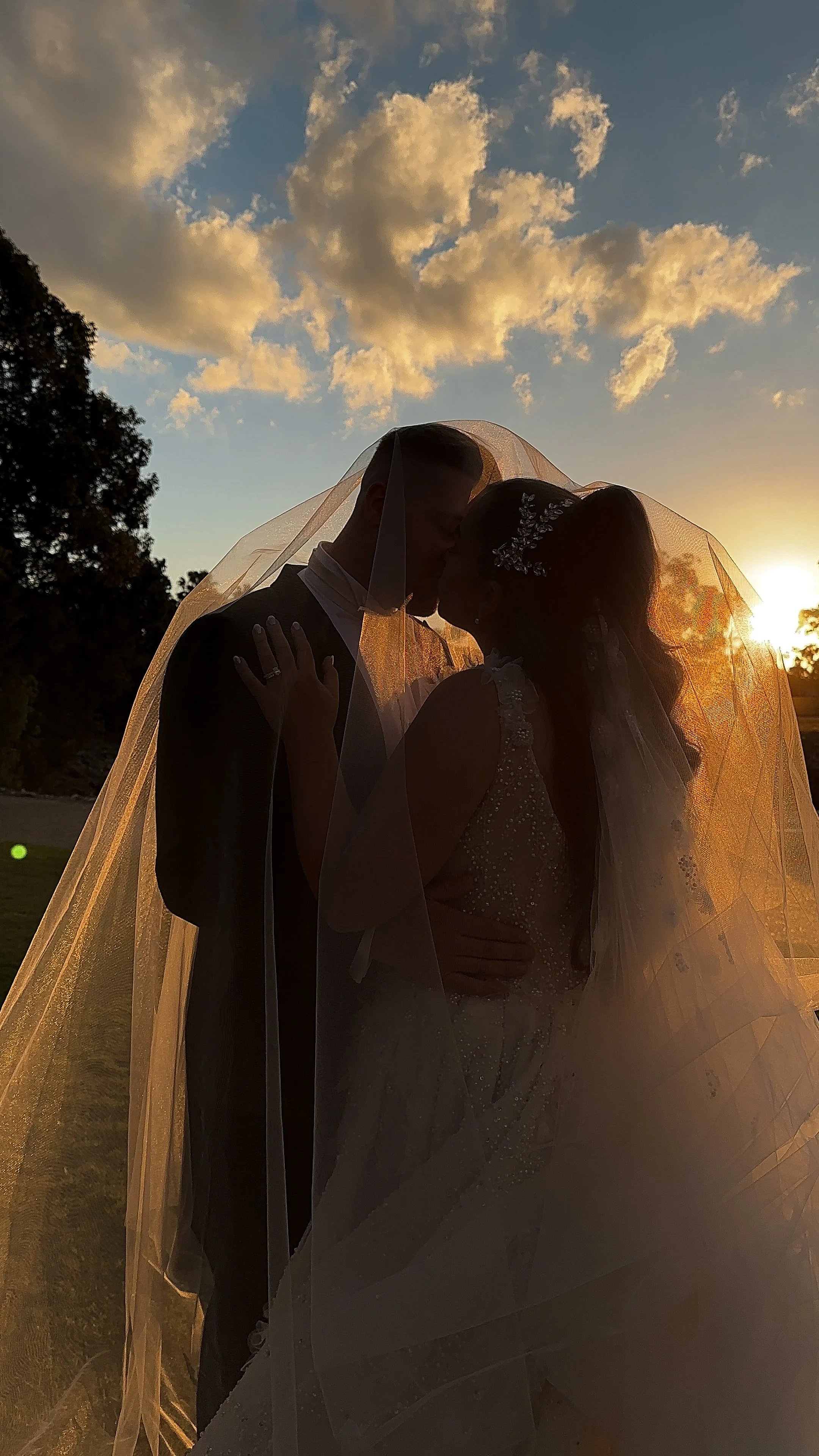 Silhouetted newlywed couple sharing an intimate moment under a veil at sunset, with clouds in a partly cloudy sky.