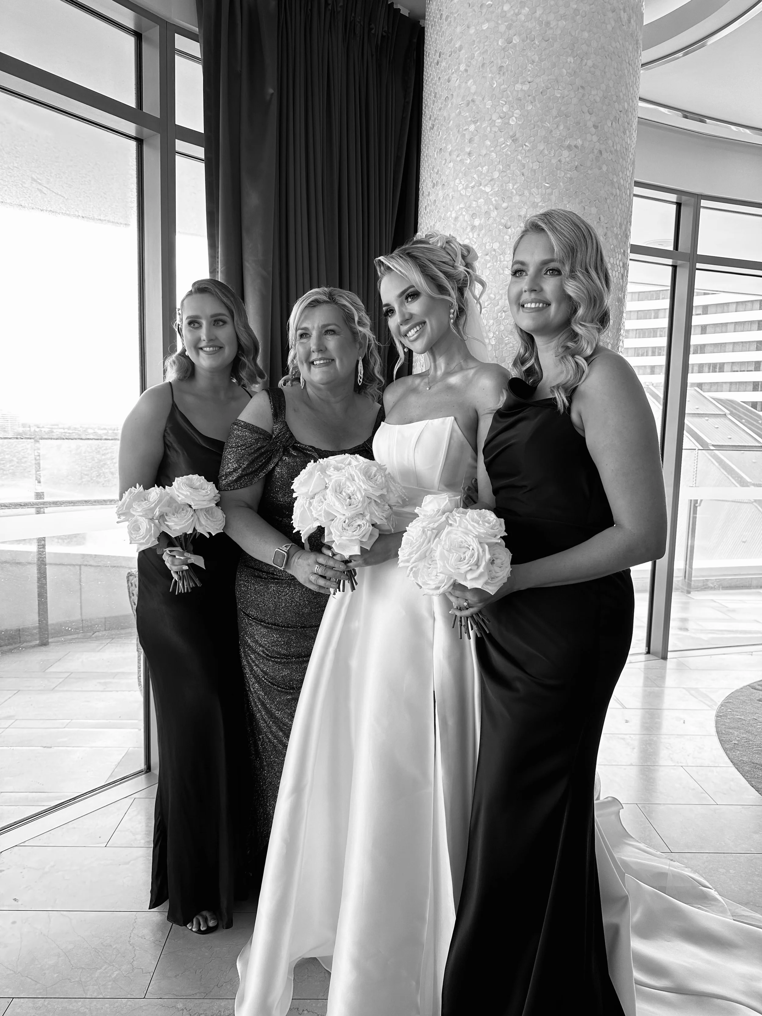 Black and white photo of a bride with three women, celebrating a wedding. The bride is in a strapless wedding gown, holding a bouquet of roses, and smiling. The women around her are dressed in formal black dresses, each holding bouquets of roses.