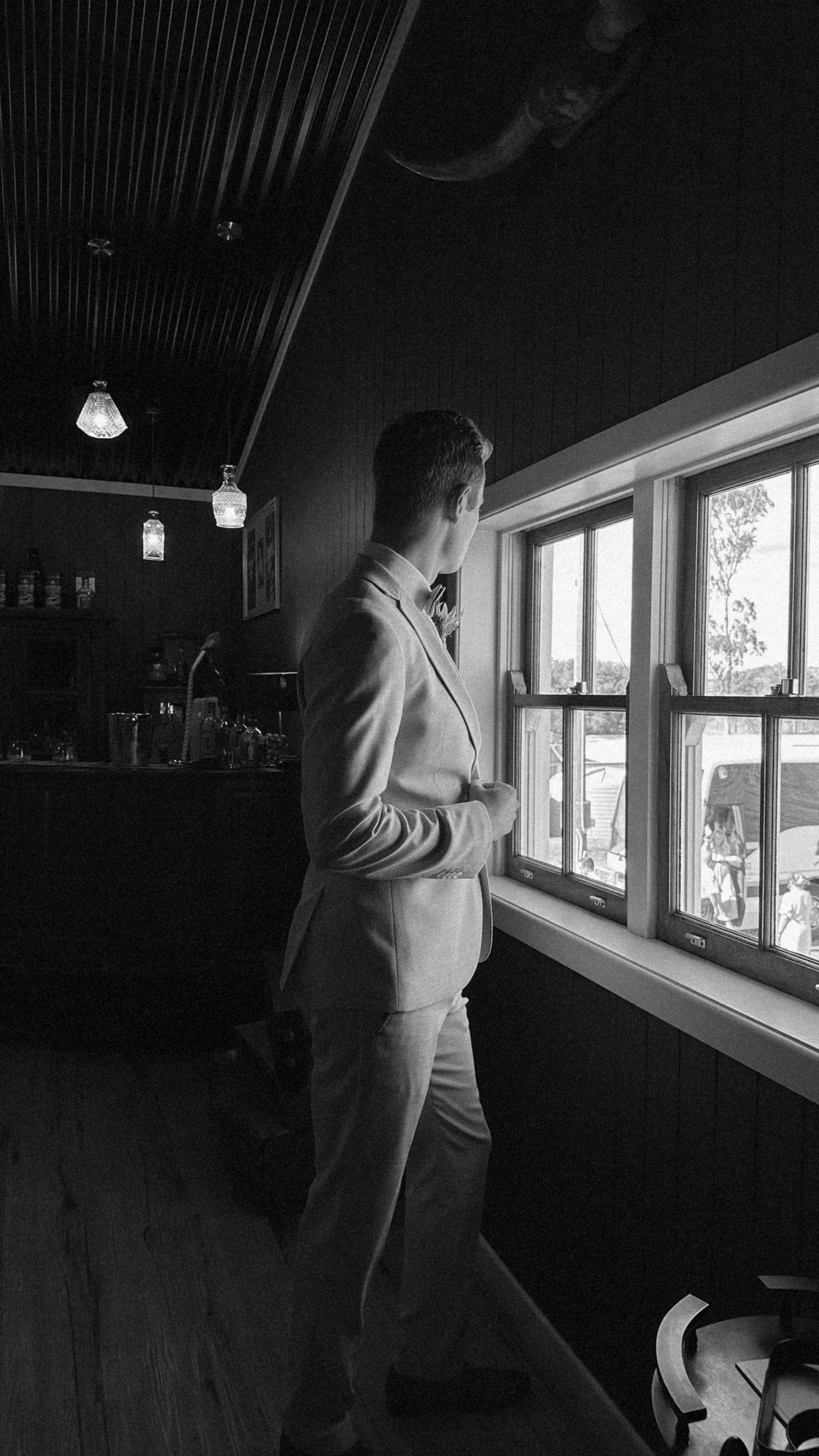 A man in a suit looking out the window inside a room, possibly a restaurant or cafe, with a bar area, hanging lights, and a view outside showing people and trees.