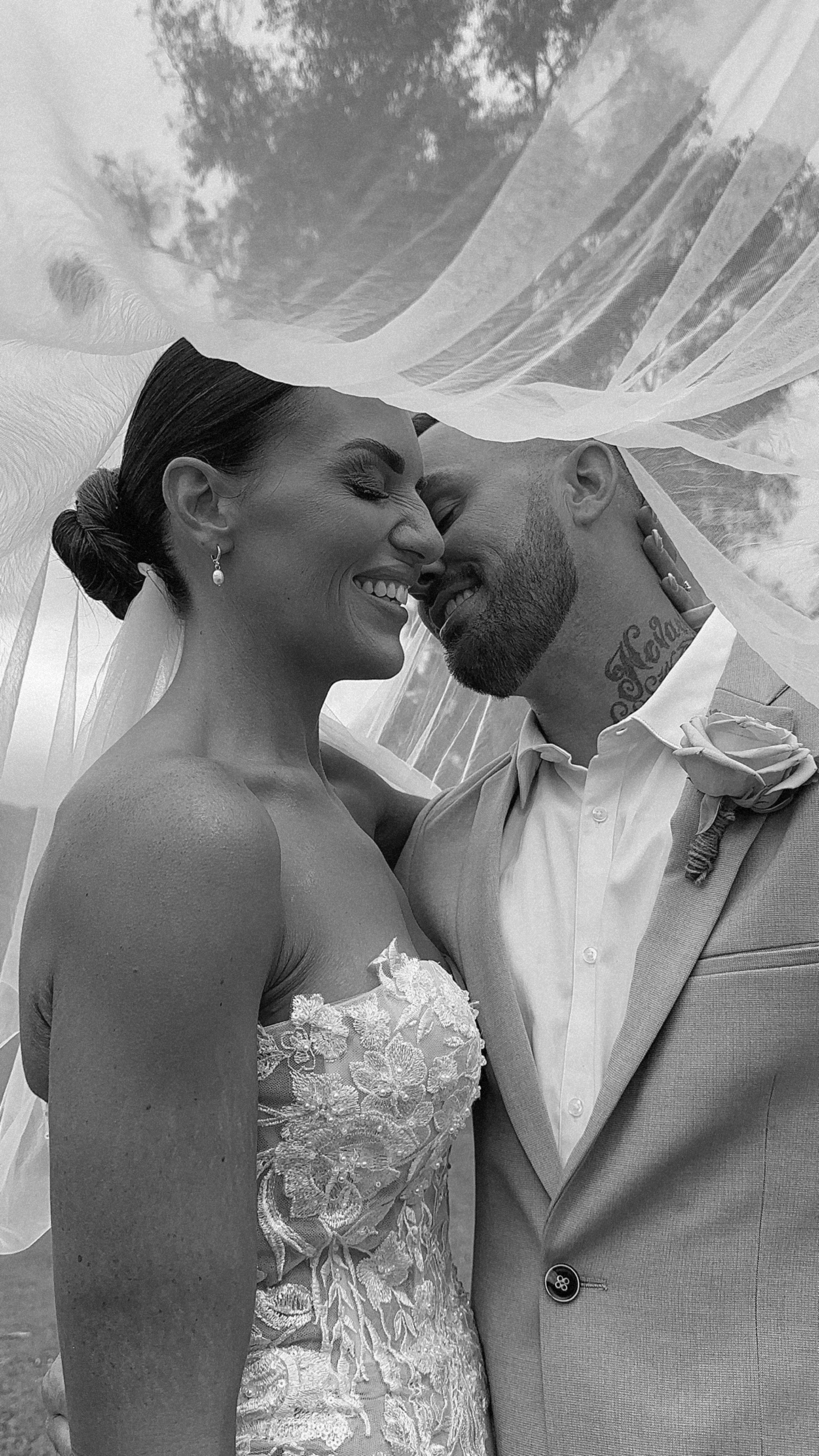 Black and white photo of a bride and groom smiling and touching faces under a sheer veil, on their wedding day.