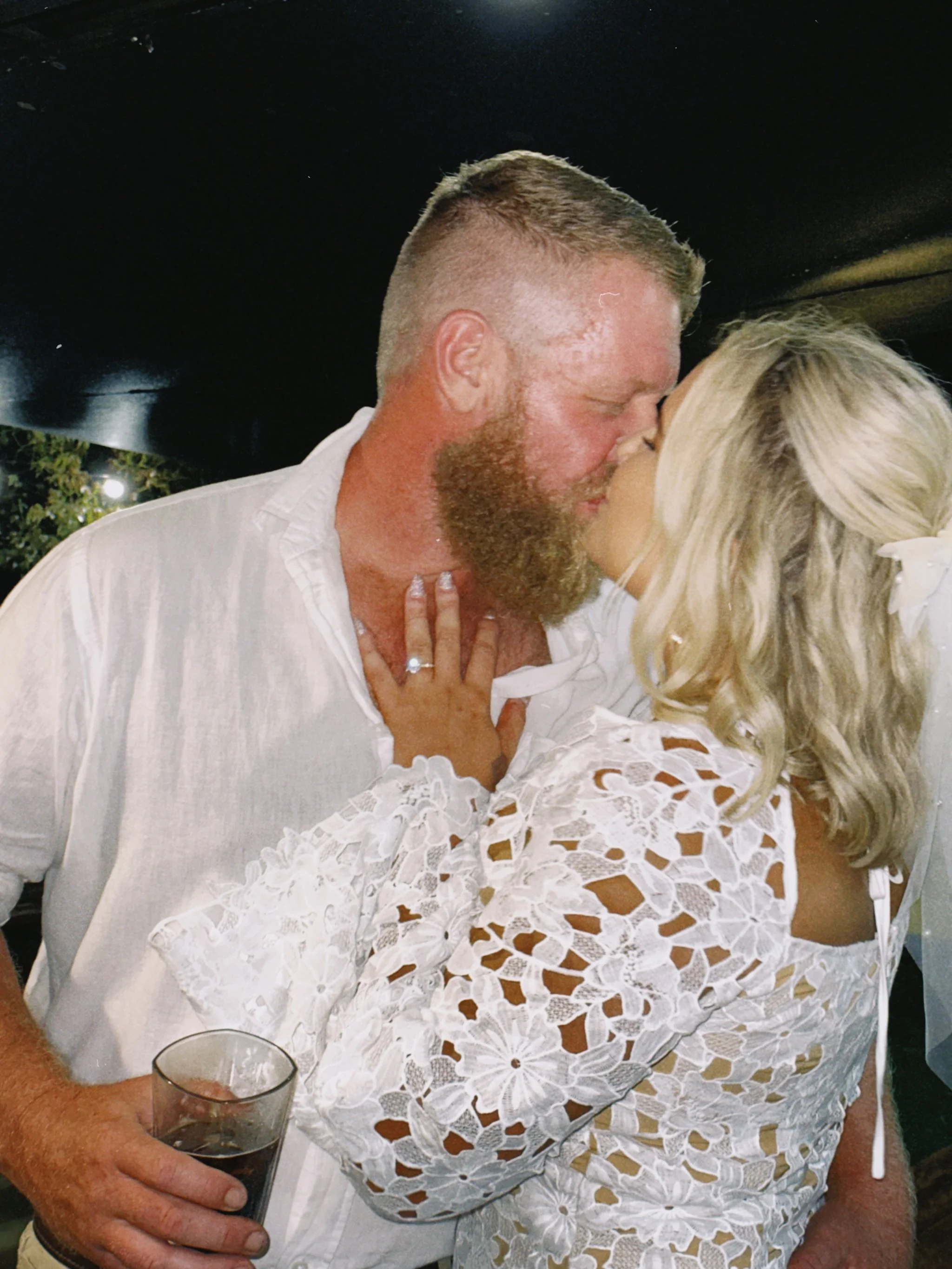 A man and woman kissing at a wedding reception, with the woman wearing a white lace dress, the man holding a drink, and the woman's hand with a wedding ring resting on the man's chest.