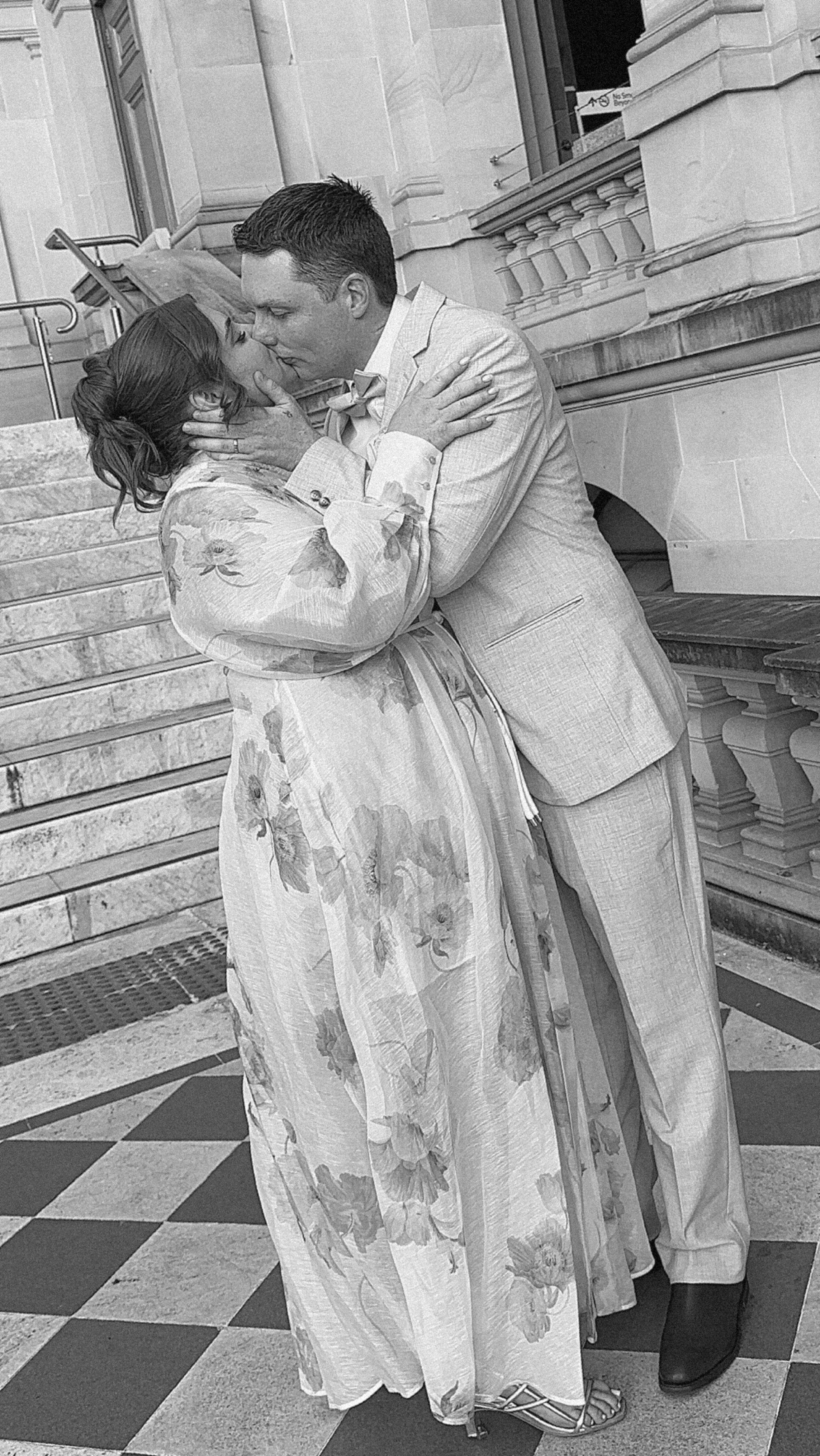 A black and white photo of a couple kissing on the steps inside a building that appears to be a courthouse or historic structure.
