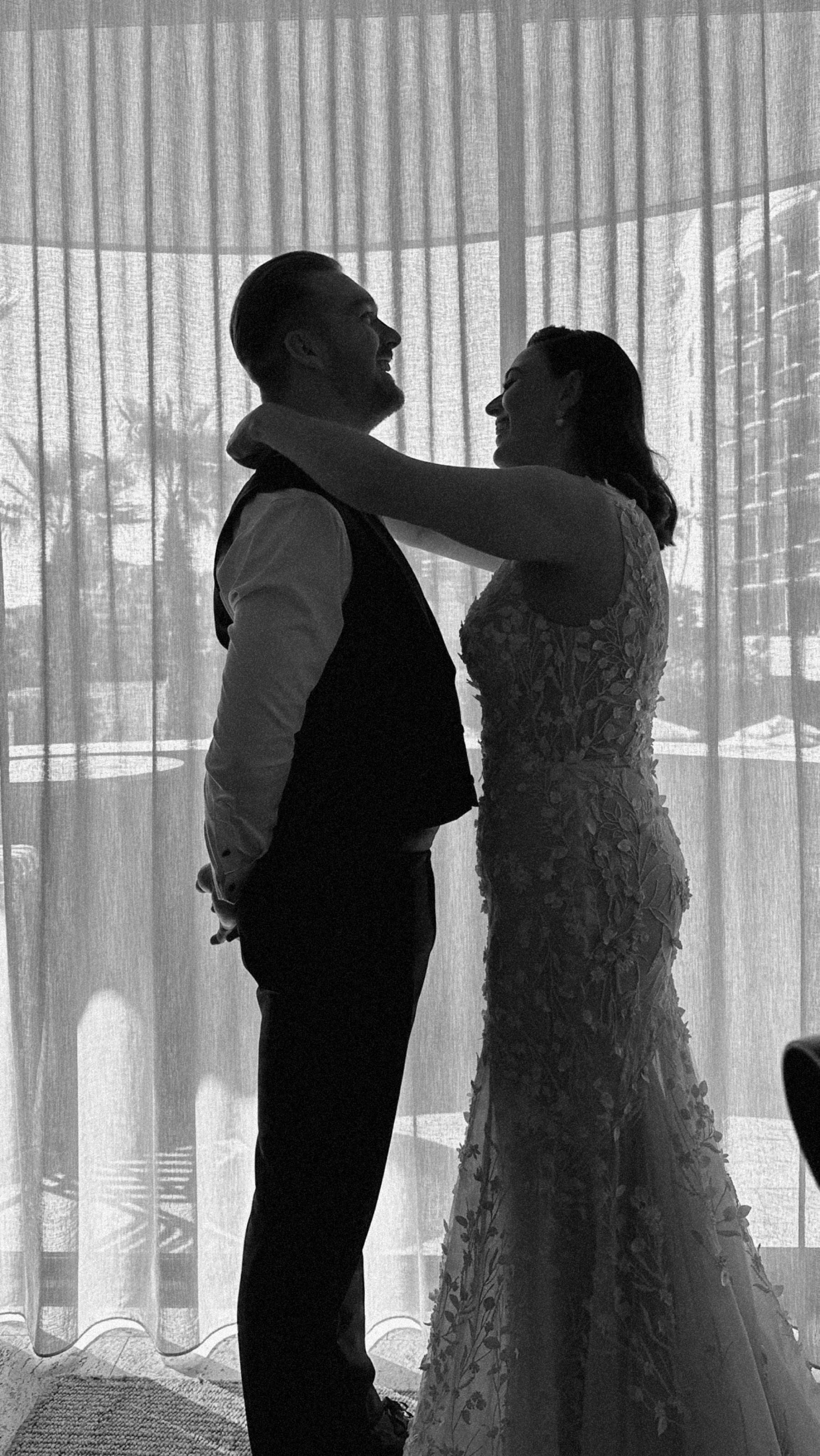 A couple in wedding attire facing each other, smiling, with a curtain and outdoor scenery in the background.