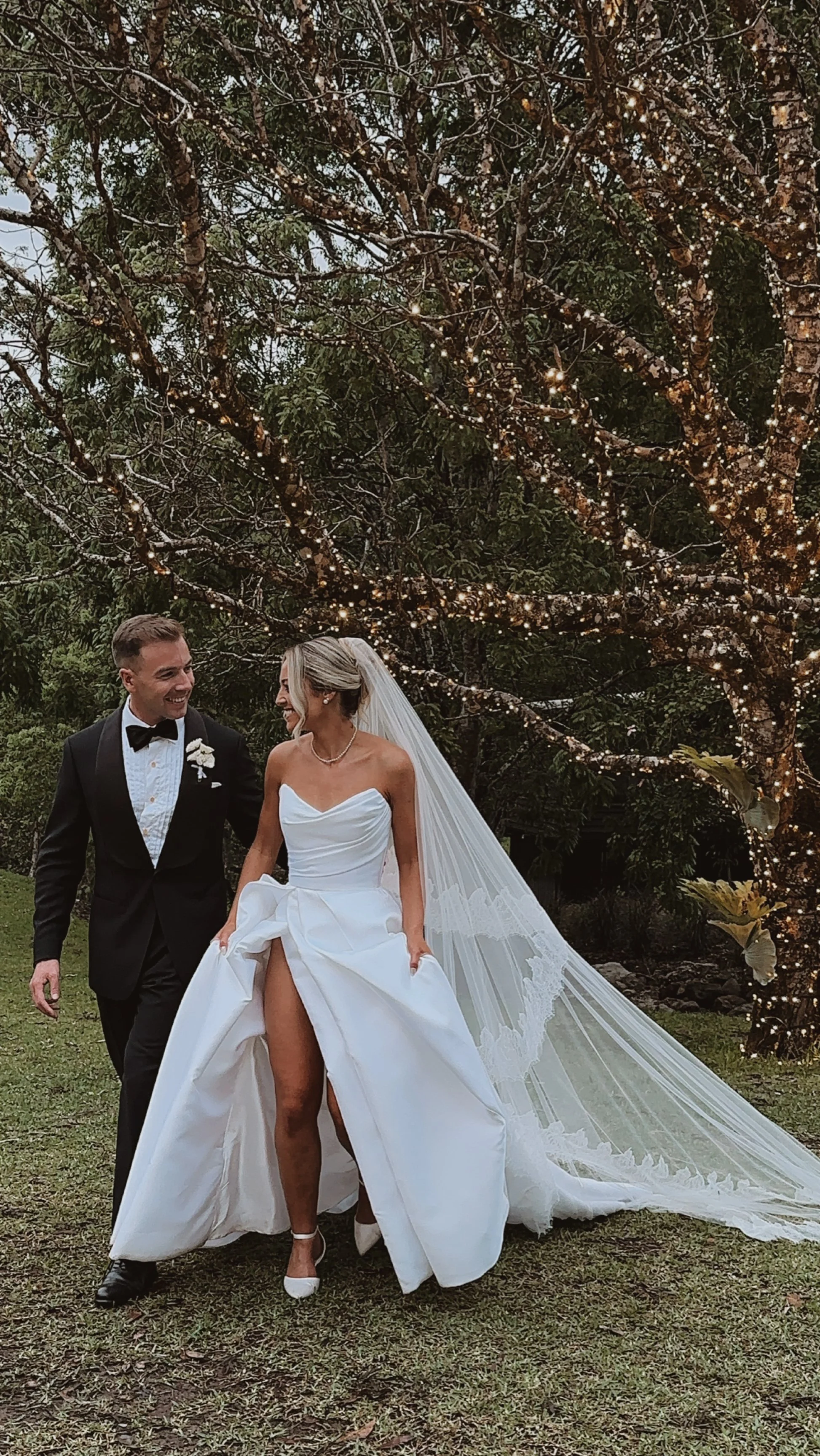 A bride and groom smiling and walking outdoors, with the bride lifting her wedding dress to show her leg, under a tree decorated with string lights.