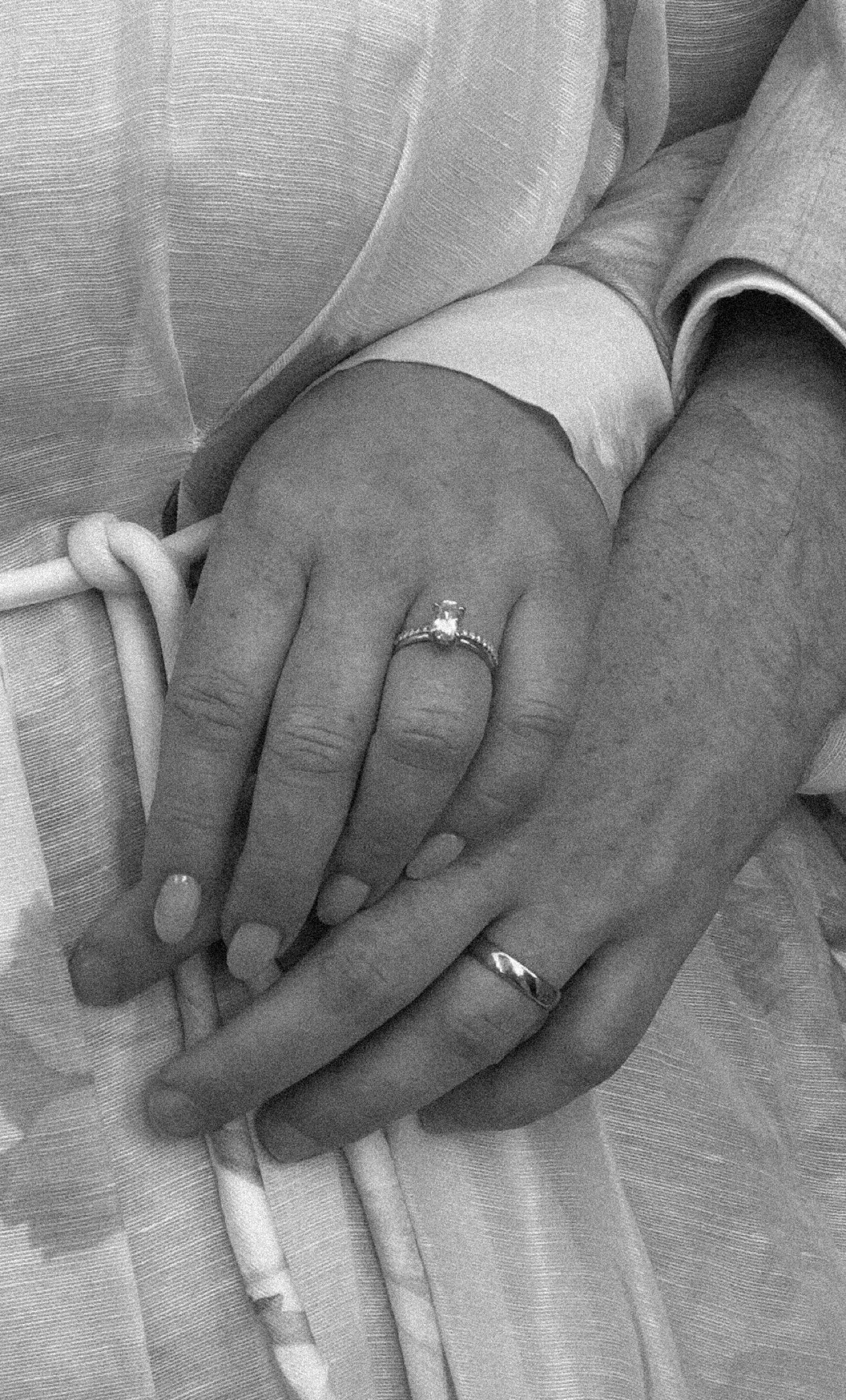 Close-up of two hands, one with an engagement ring, resting on a medical or hospital bed with medical tubes nearby.
