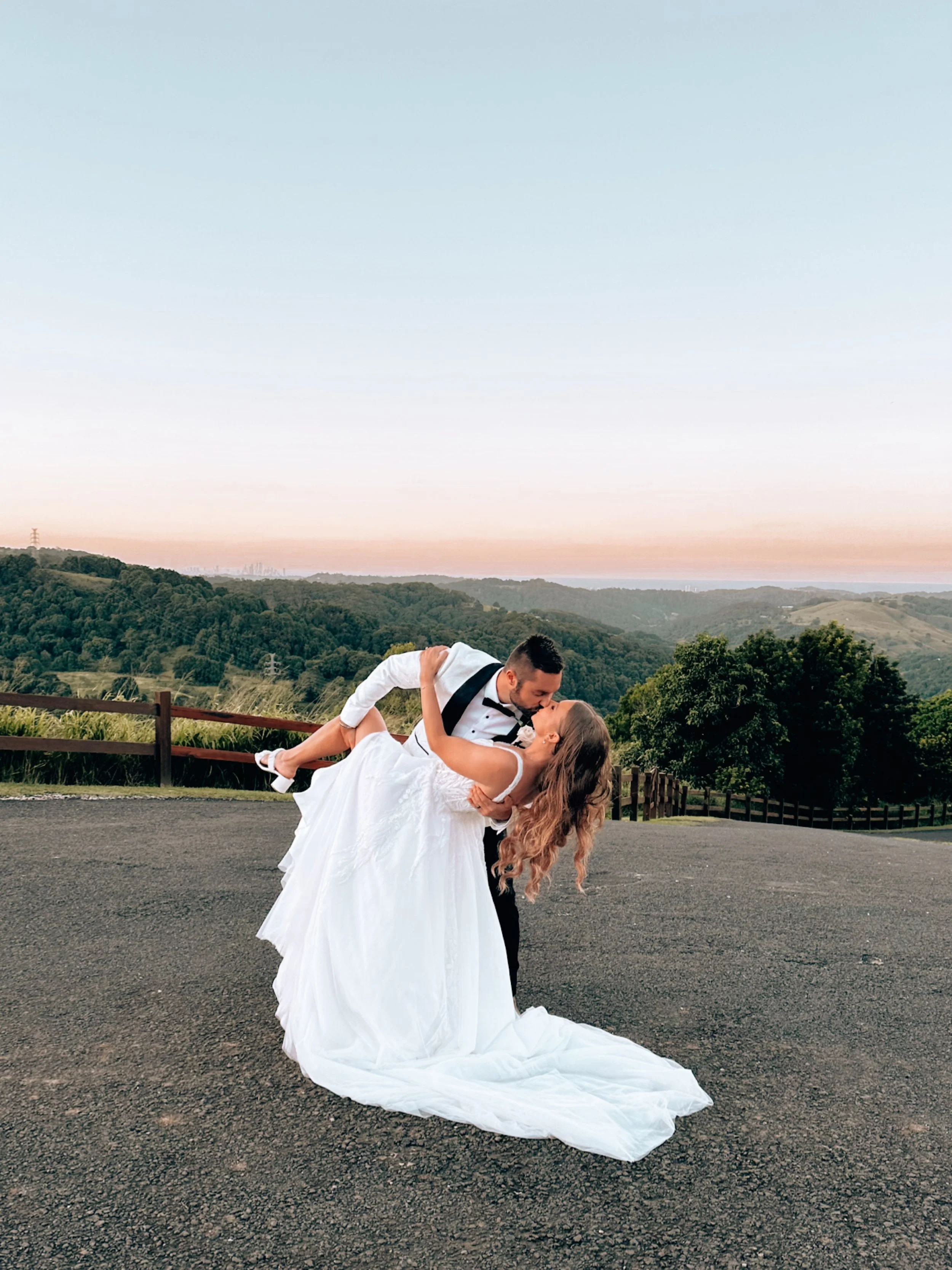 A newlywed couple dancing outdoors on a paved road with scenic hills and trees in the background at dusk. The groom is dipping the bride, who is wearing a white wedding dress and the groom is in a tuxedo.
