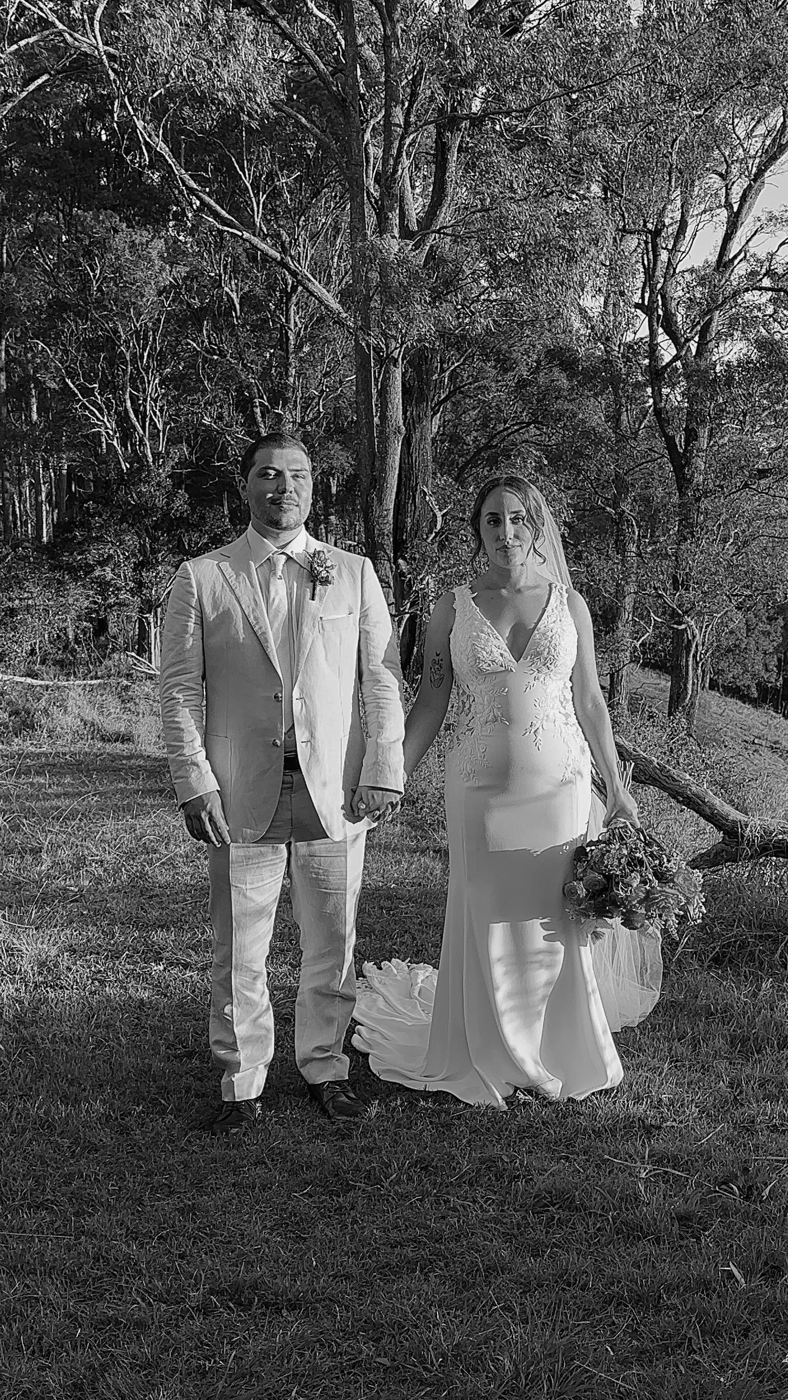 Black and white photo of a bride and groom standing outdoors in a forested area, holding hands, with the bride holding a bouquet of flowers.