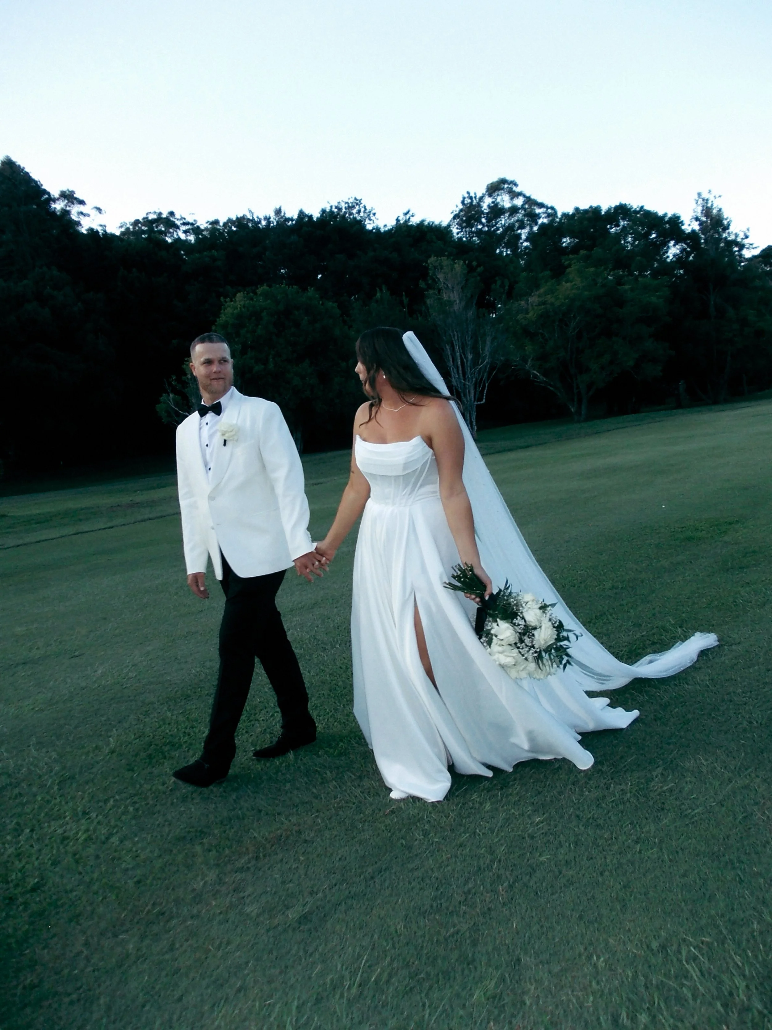 A bride and groom holding hands on a grassy field, with trees in the background, during their wedding.