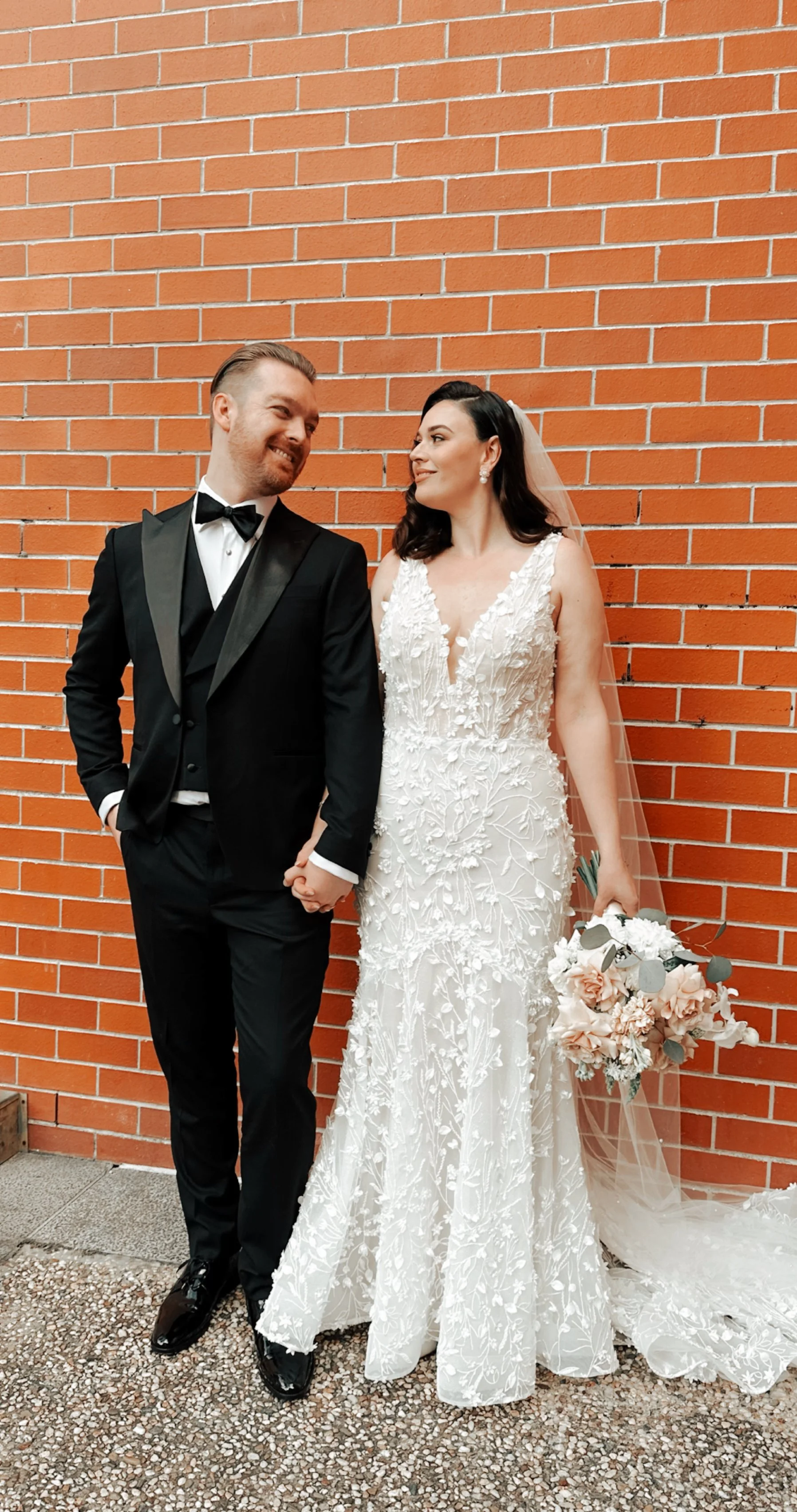 A bride and groom holding hands and smiling at each other, standing against a red brick wall. The bride wears a white lace wedding dress with a veil, holding a bouquet of white and blush flowers. The groom wears a black tuxedo with a black bow tie.