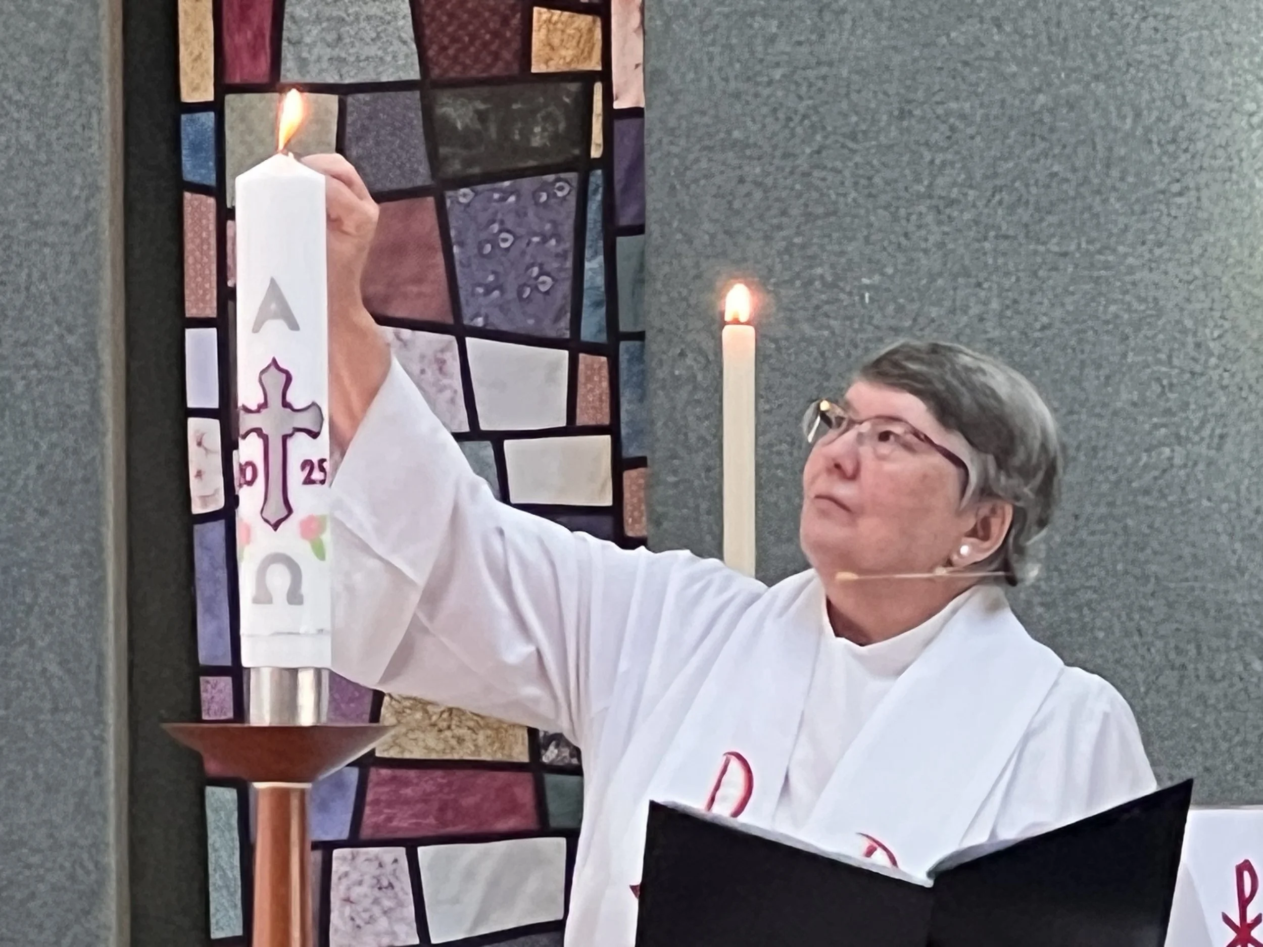 A woman in white liturgical vestments lighting a tall candle with a flame, in front of a stained glass window with various colored panes.