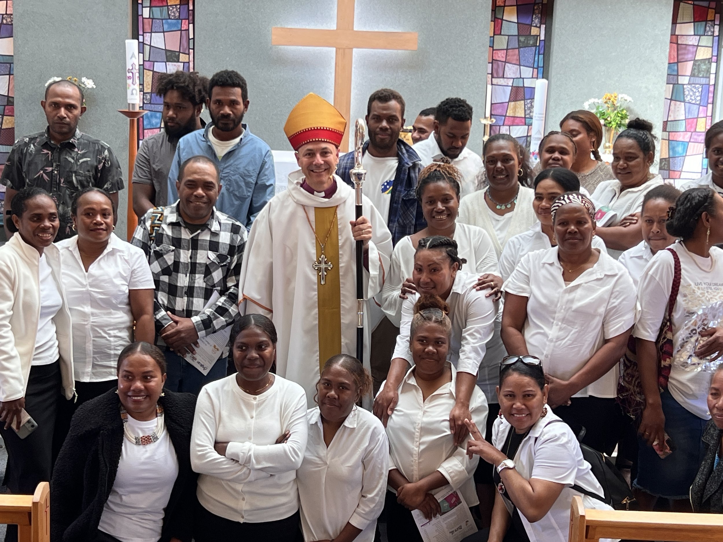 Group of diverse people gathered in a church with stained glass windows, featuring a bishop in white robes and a red mitre, holding a staff, surrounded by smiling individuals in white shirts and casual attire.
