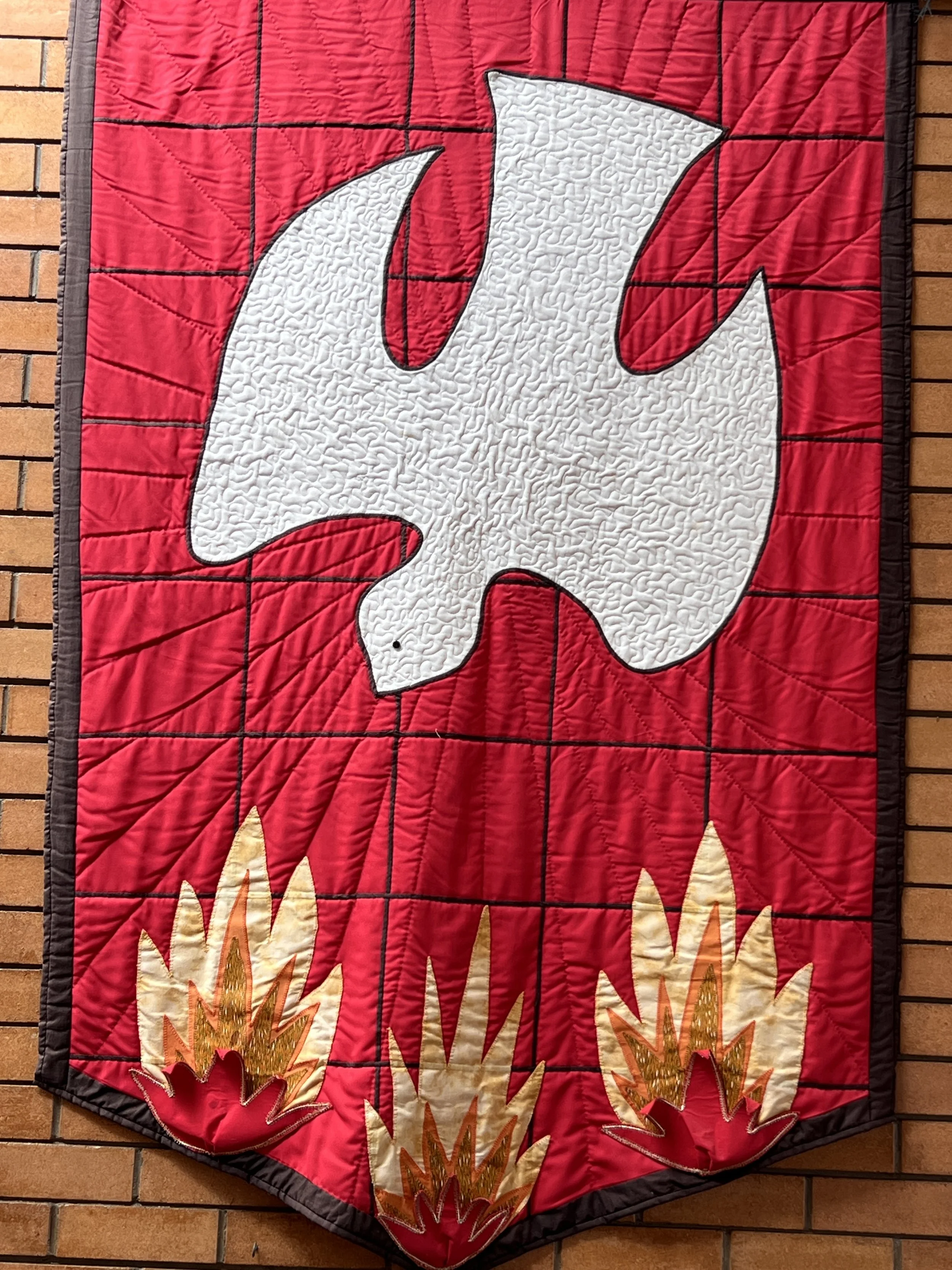 Decorative quilt with a white dove flying above stylized flames on a red background, hung on a brick wall.