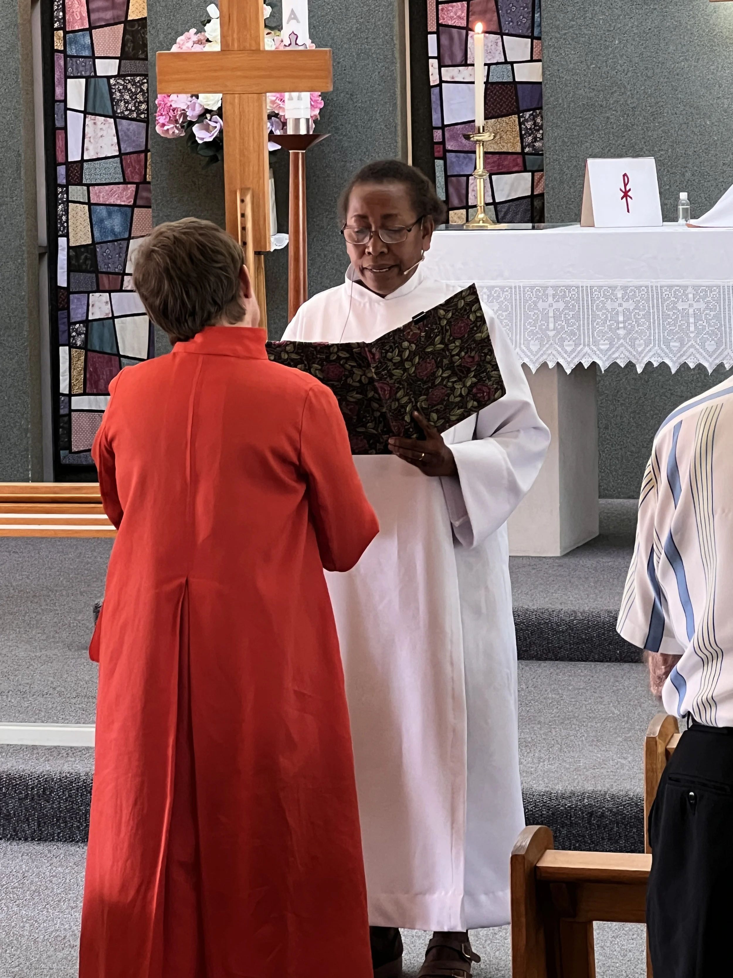 A woman in white clergy robes and glasses reads from a book during a church service, facing a woman in a red dress. The altar behind them has a large wooden cross, lit candle, and stained glass windows.